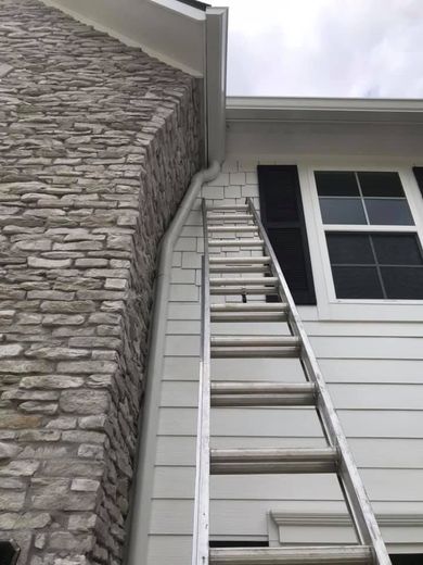 Ladder leaning against a house, near a stone chimney and white siding, for gutter work.
