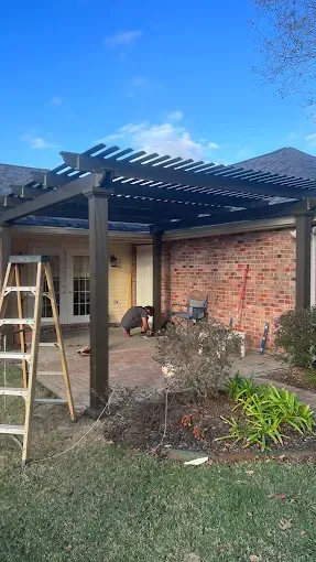 A dark-colored pergola over a brick patio with a ladder, plants, and a house, against a blue sky.