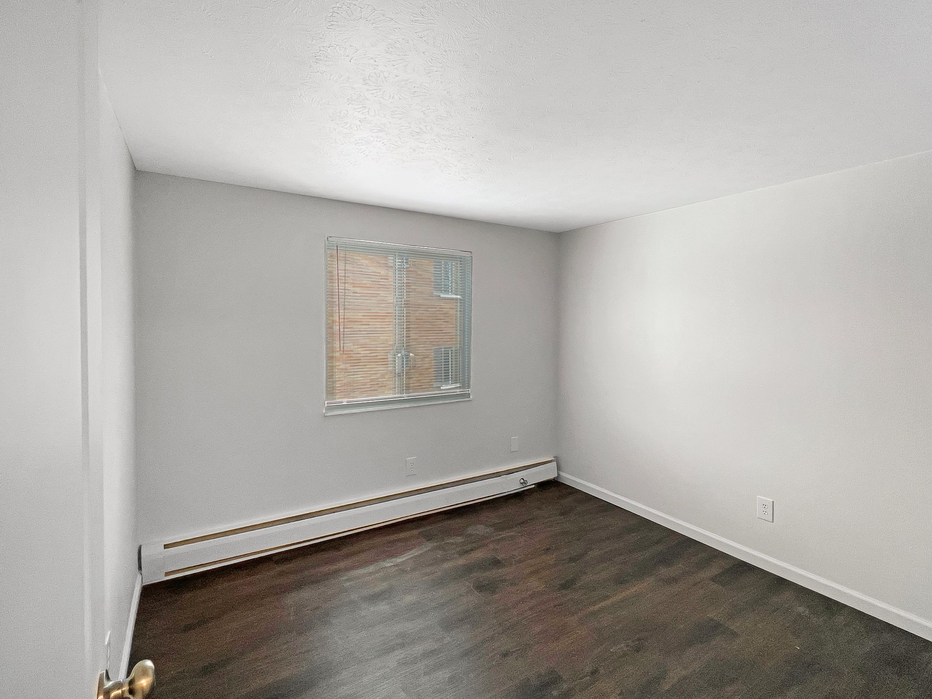 Empty room with dark hardwood floors, light gray walls, window with blinds.
