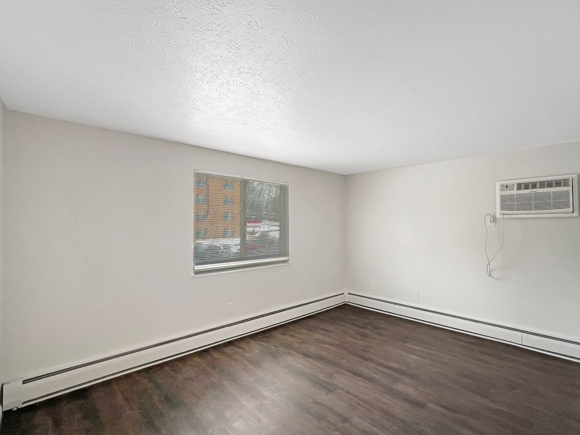 Empty room with dark wood flooring, white walls and ceiling, window, and air conditioner.