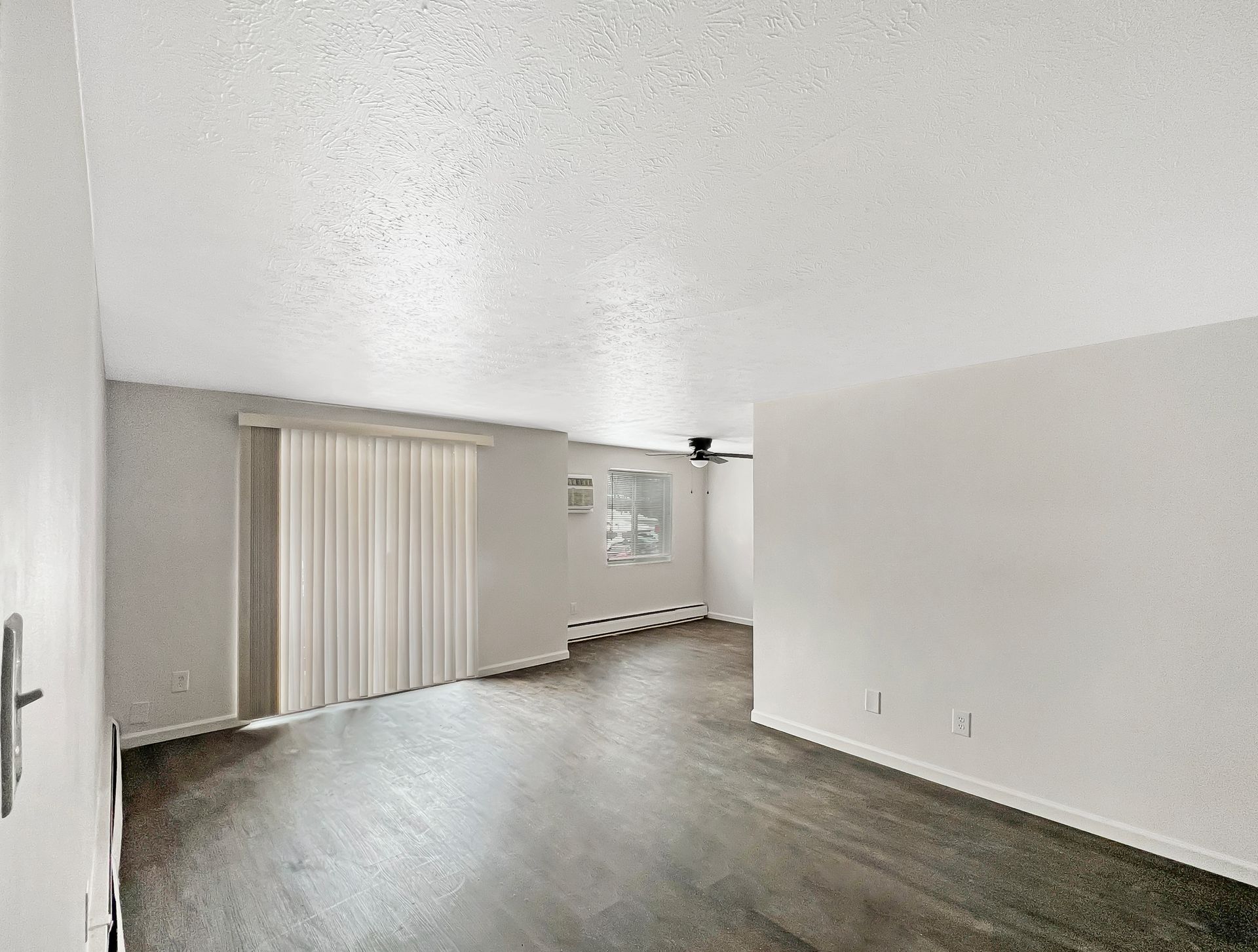 Empty living room with dark wood flooring, white walls and ceiling, and sliding glass door with a cream colored curtain.