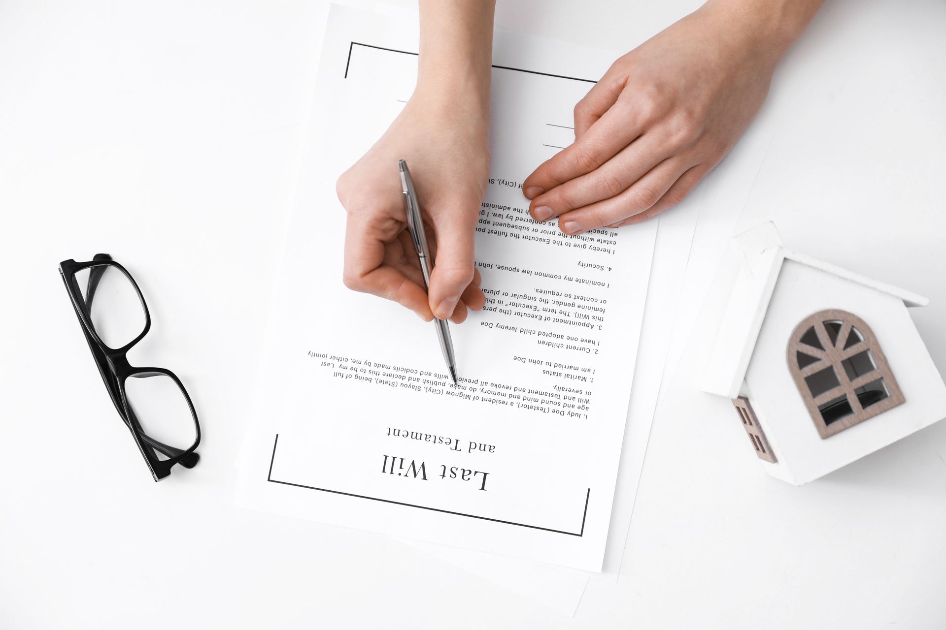 Person signing a last will document beside eyeglasses and a small model house.