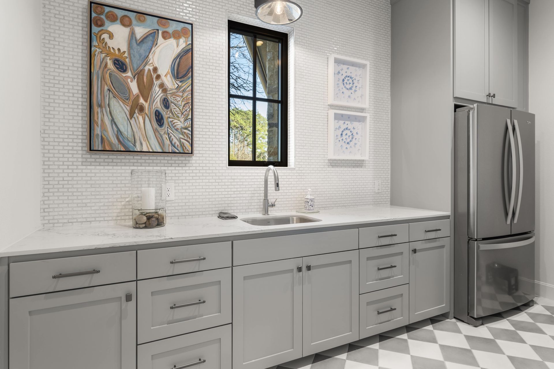 A laundry room with gray cabinets , a stainless steel refrigerator , a sink and a window.
