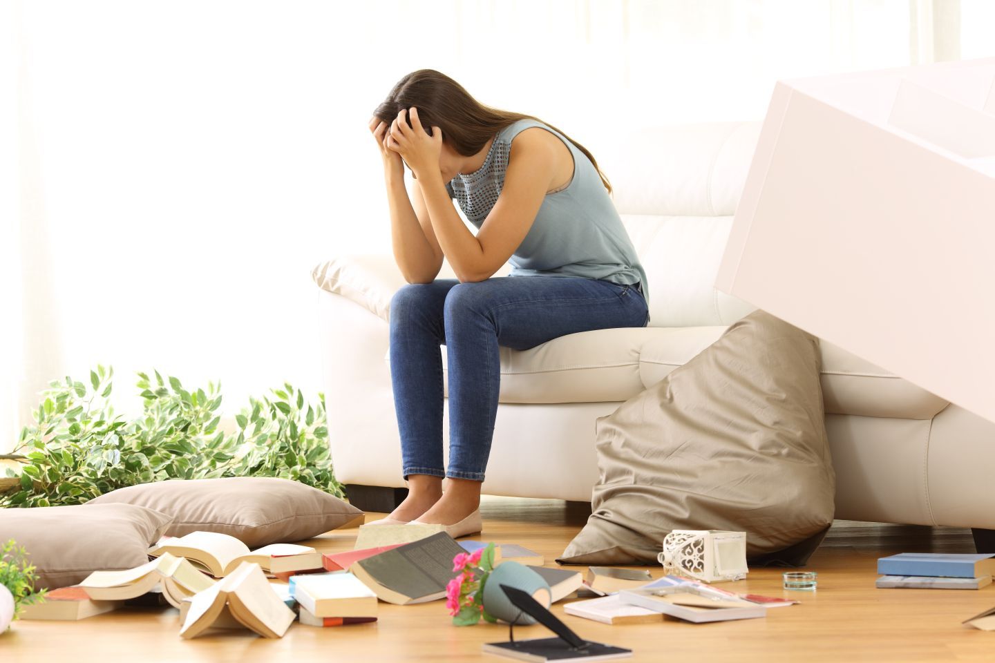 Woman sitting on couch, head in hands, amidst scattered objects in a messy living room.