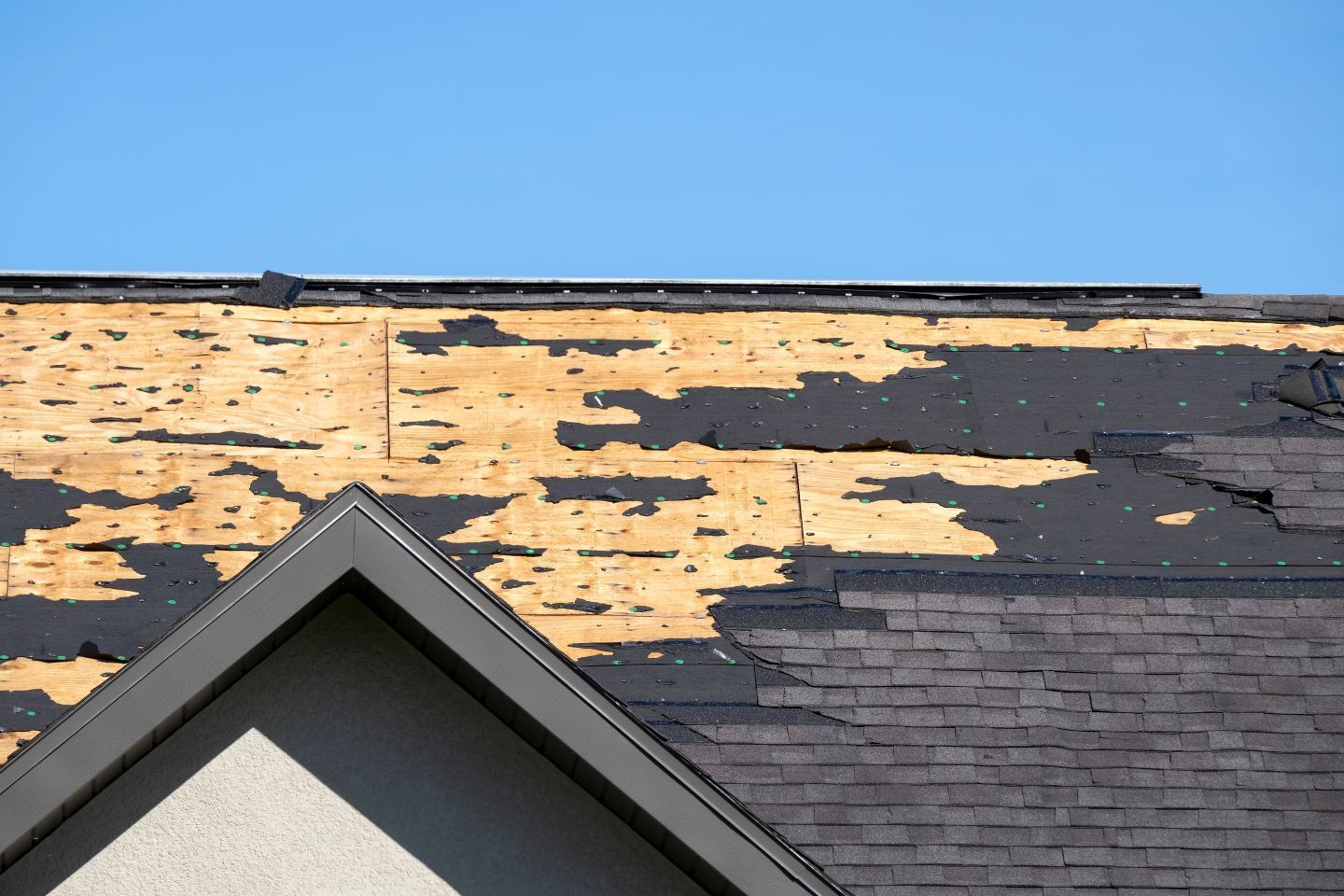 Damaged asphalt shingles on a house roof, with exposed wood decking against a clear blue sky.