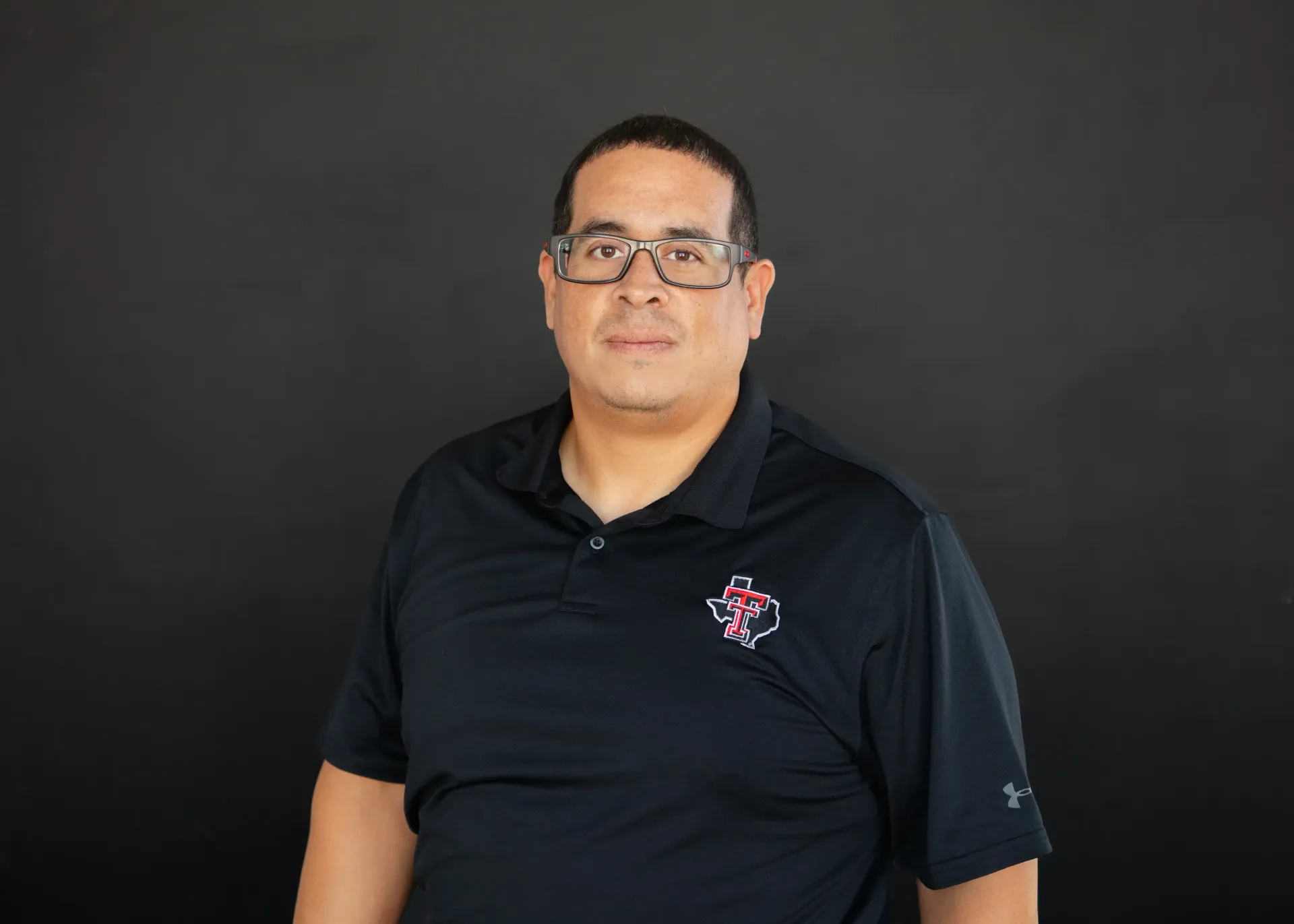 Man wearing glasses and a black polo shirt, standing in front of a dark background.