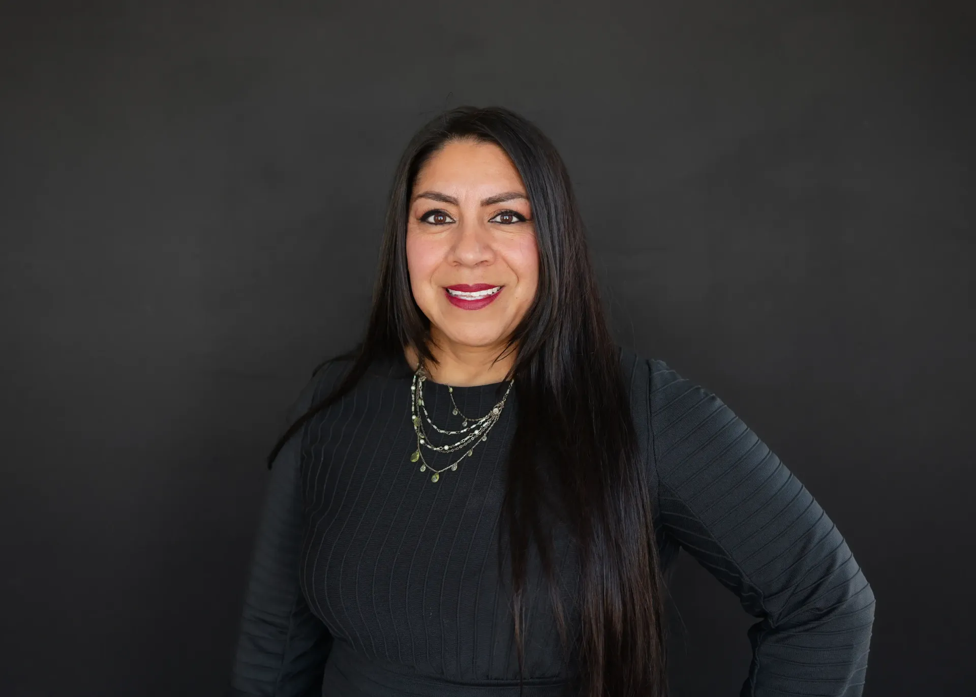 Woman with long dark hair smiles in front of a black background, wearing a dark sweater and necklace.
