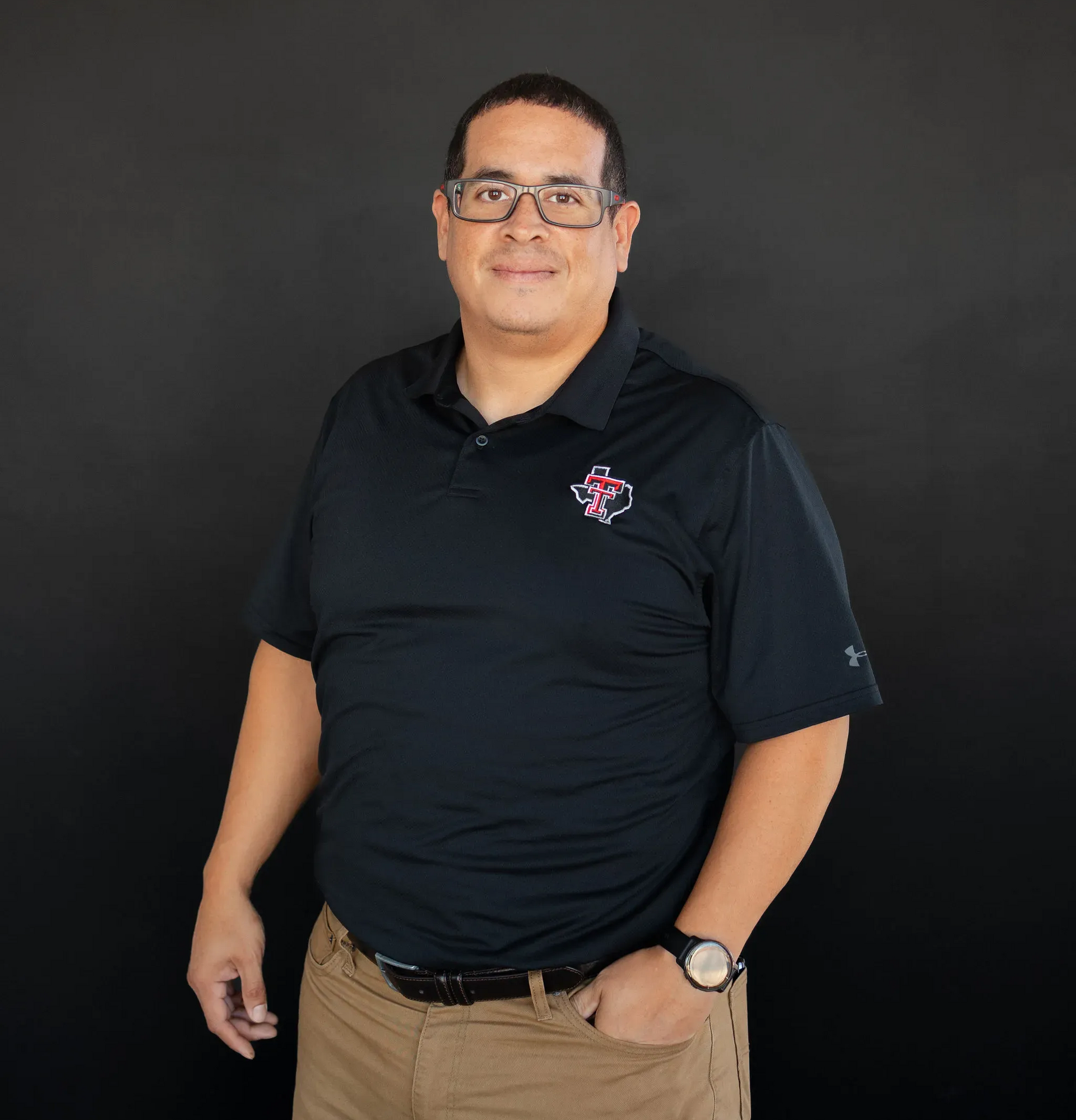 Man wearing glasses, black polo shirt, khaki pants, hands in pockets, standing against a black backdrop.