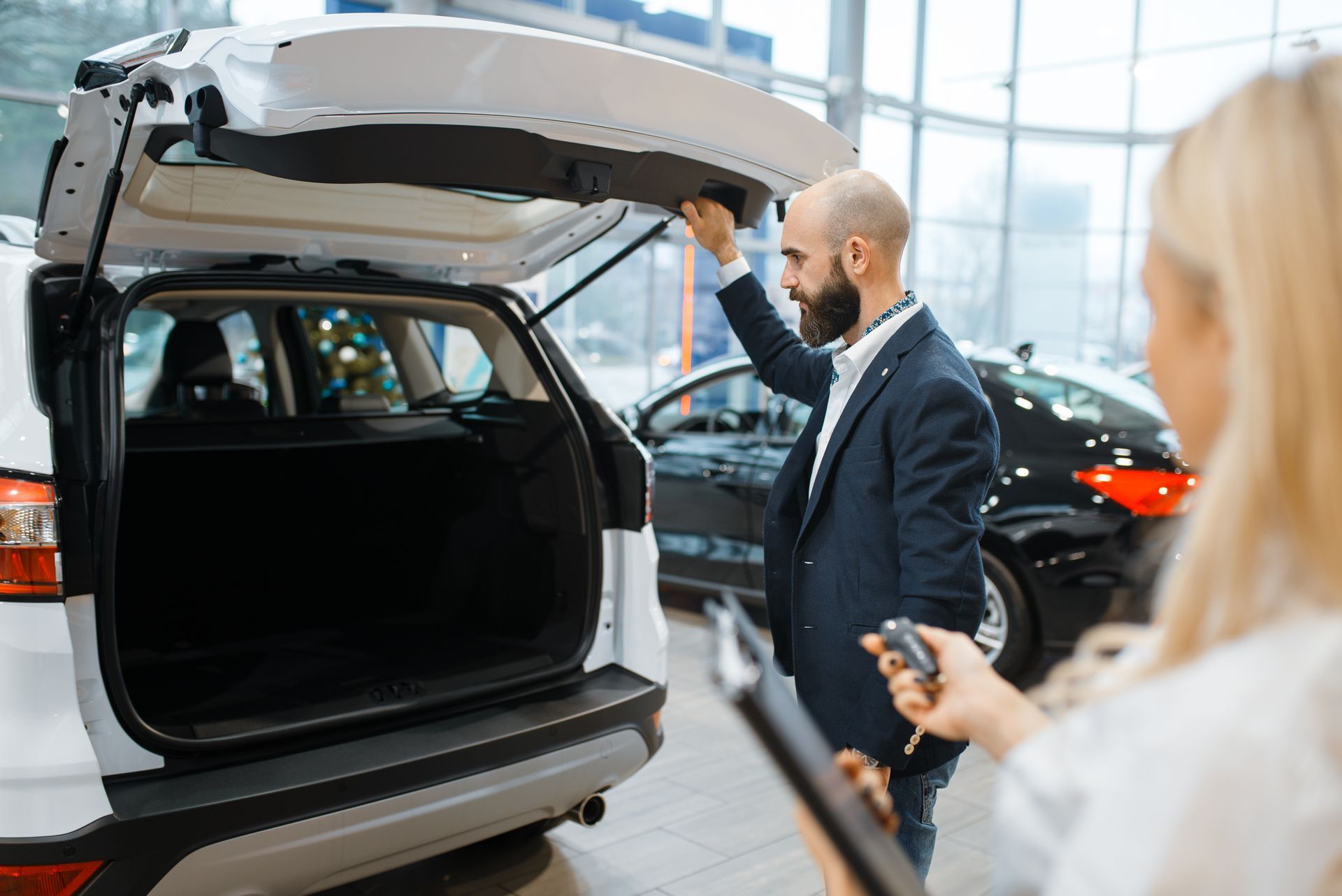 A sales consultant shows a vehicle's open trunk to a customer in a bright car dealership showroom.