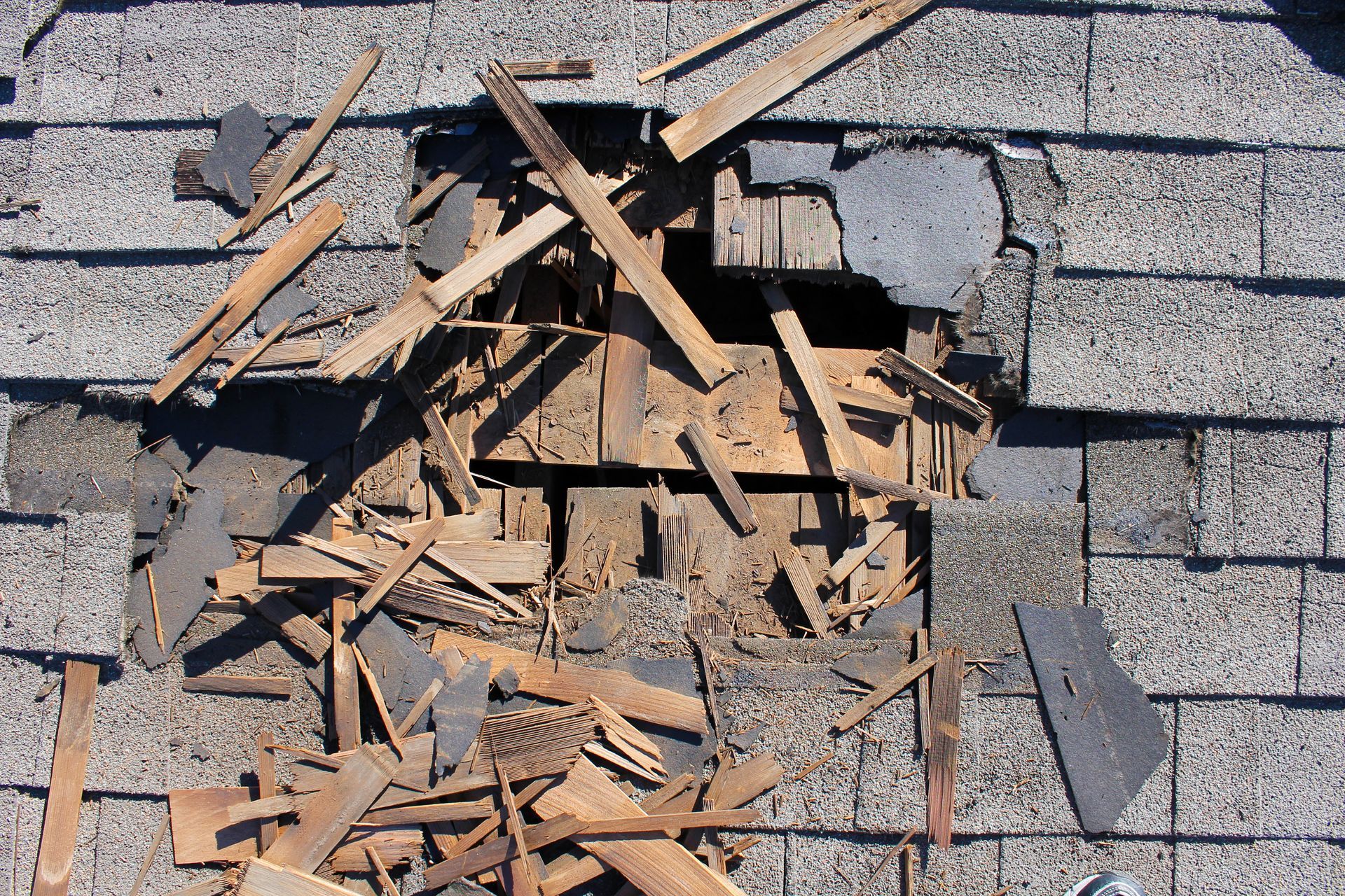 A damaged gray asphalt shingle roof with a large hole revealing broken wooden roof deck boards and debris.