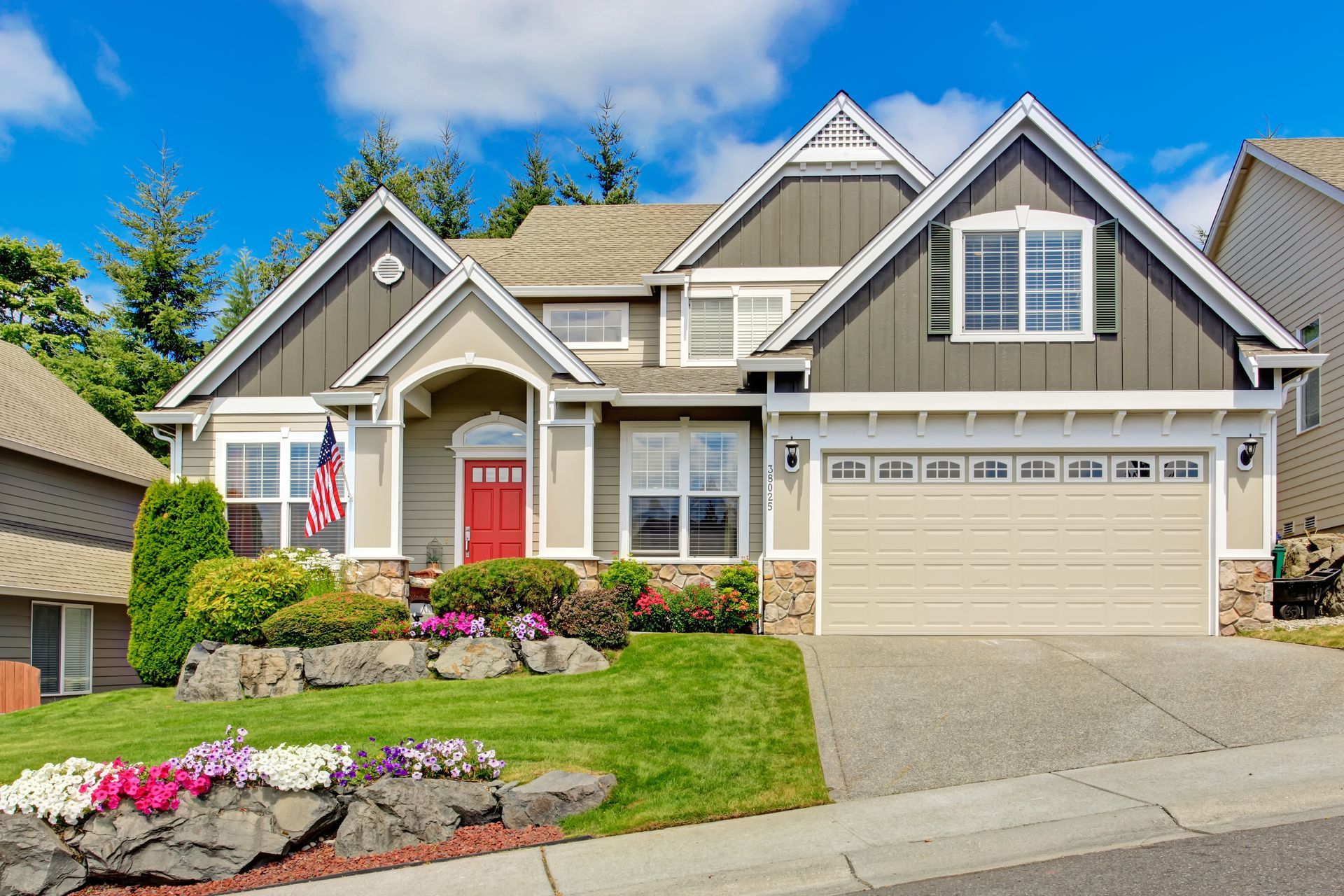 A two-story suburban home with tan siding, a red front door, a two-car garage, and a landscaped yard with flowers.