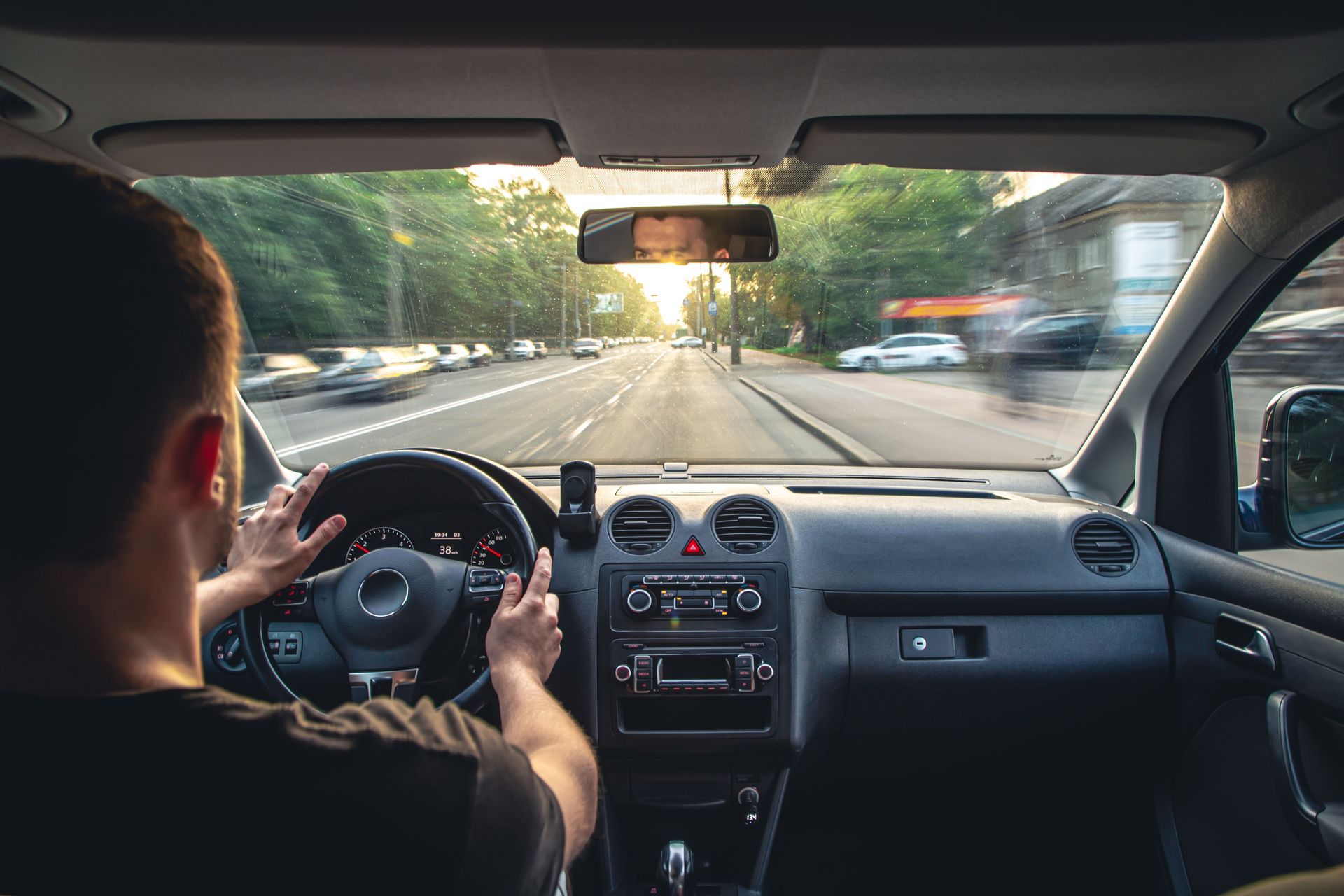 A person driving a car on a road during the day, with bright sunlight visible through the windshield.