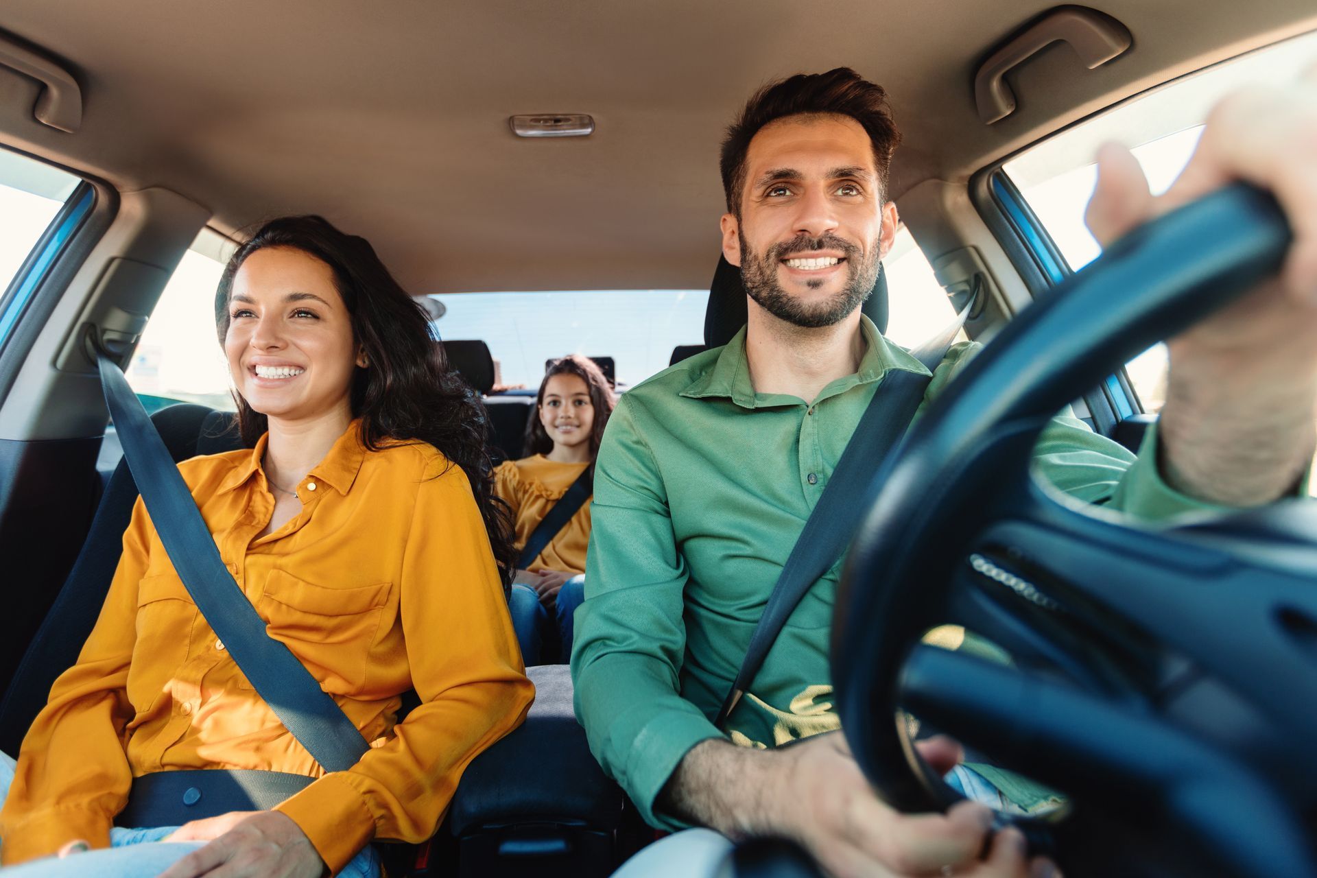 A family of three smiling while riding in a car, with the driver steering and a passenger seated in the back.