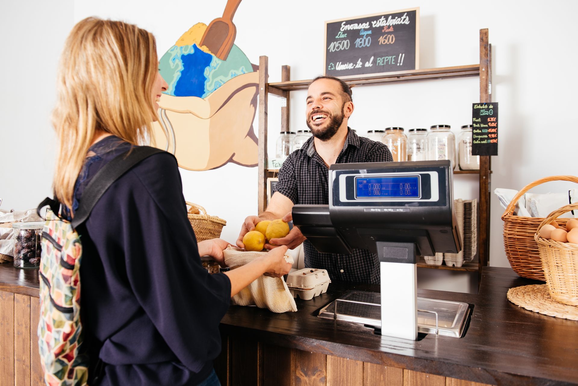 A customer hands a paper bag of produce to a smiling clerk at a shop counter with a digital scale.