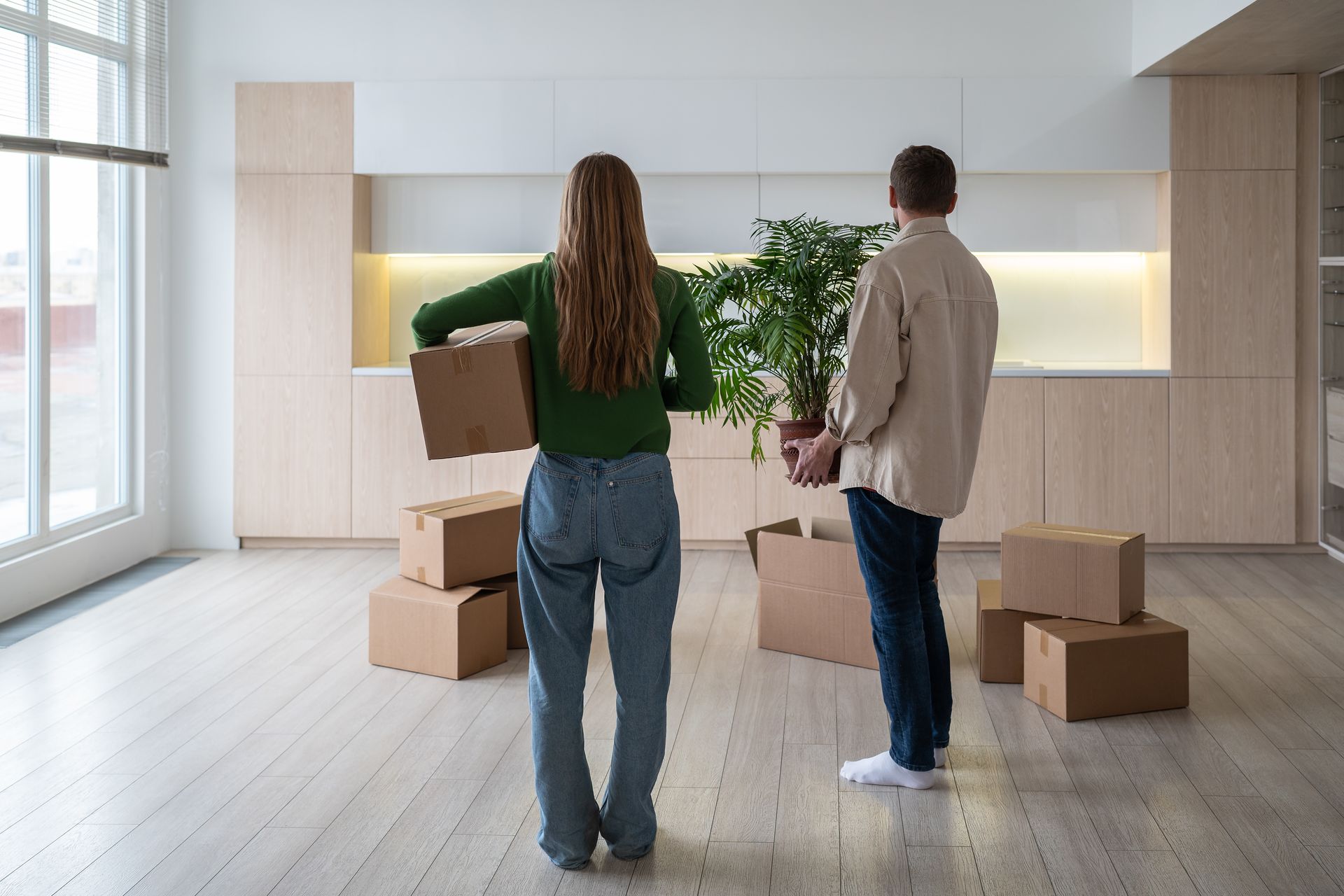 Two people carrying moving boxes and a potted plant stand in a light-filled room with wooden cabinets and flooring.