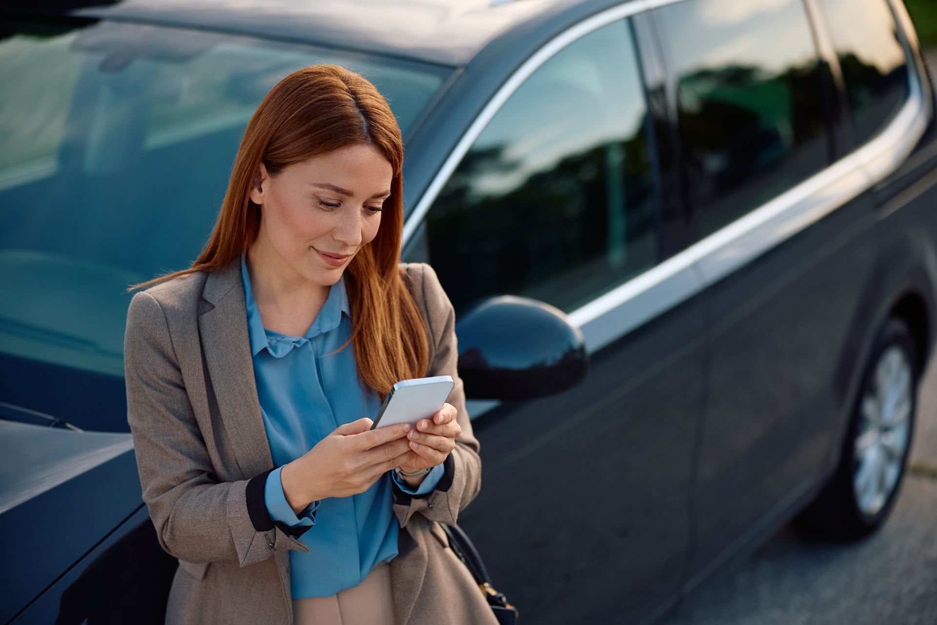 A professional wearing a blazer stands by a dark car, smiling while looking at a smartphone held in both hands.