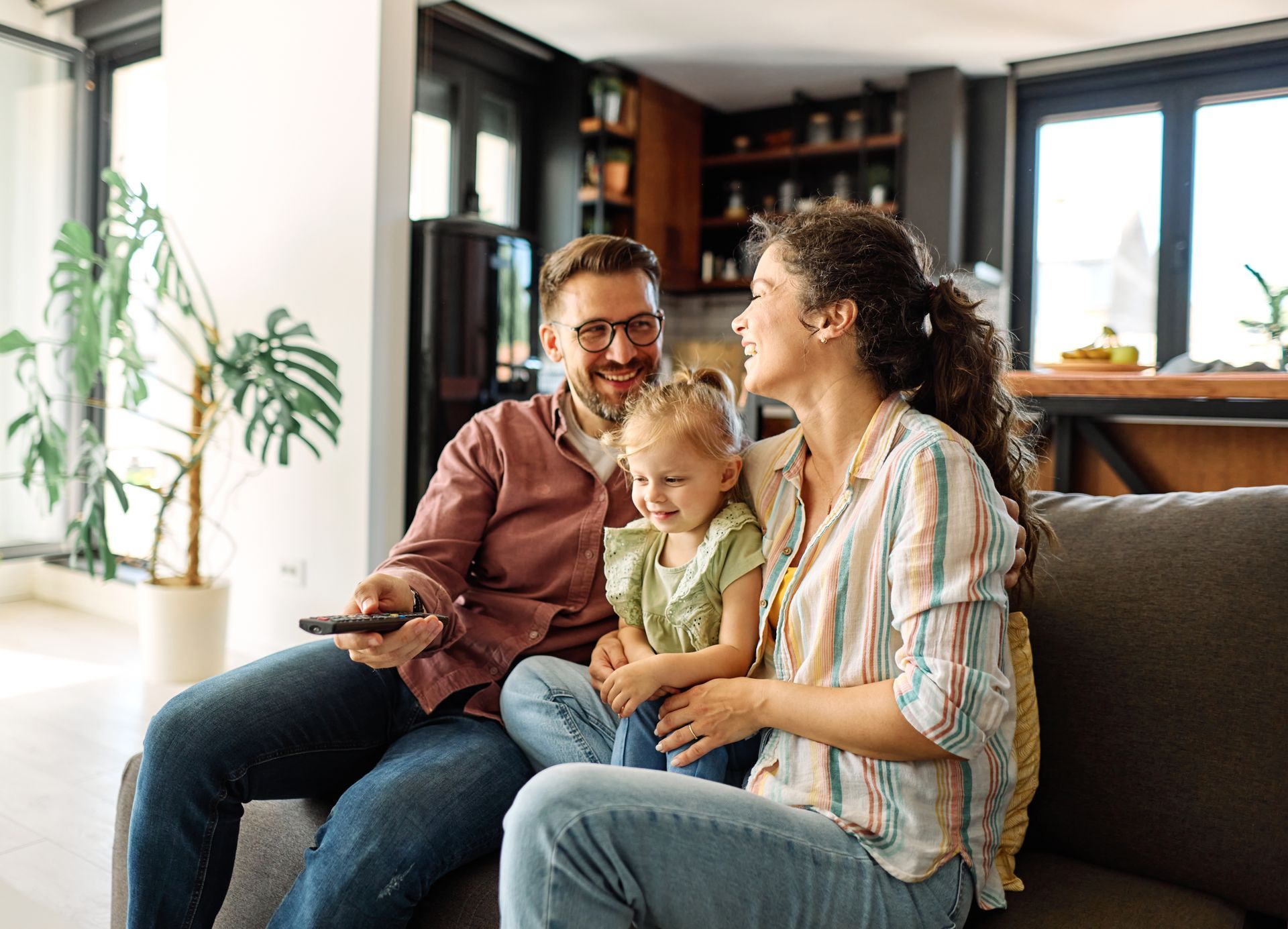 A family sitting on a sofa in a living room, smiling at each other while one person holds a remote control.