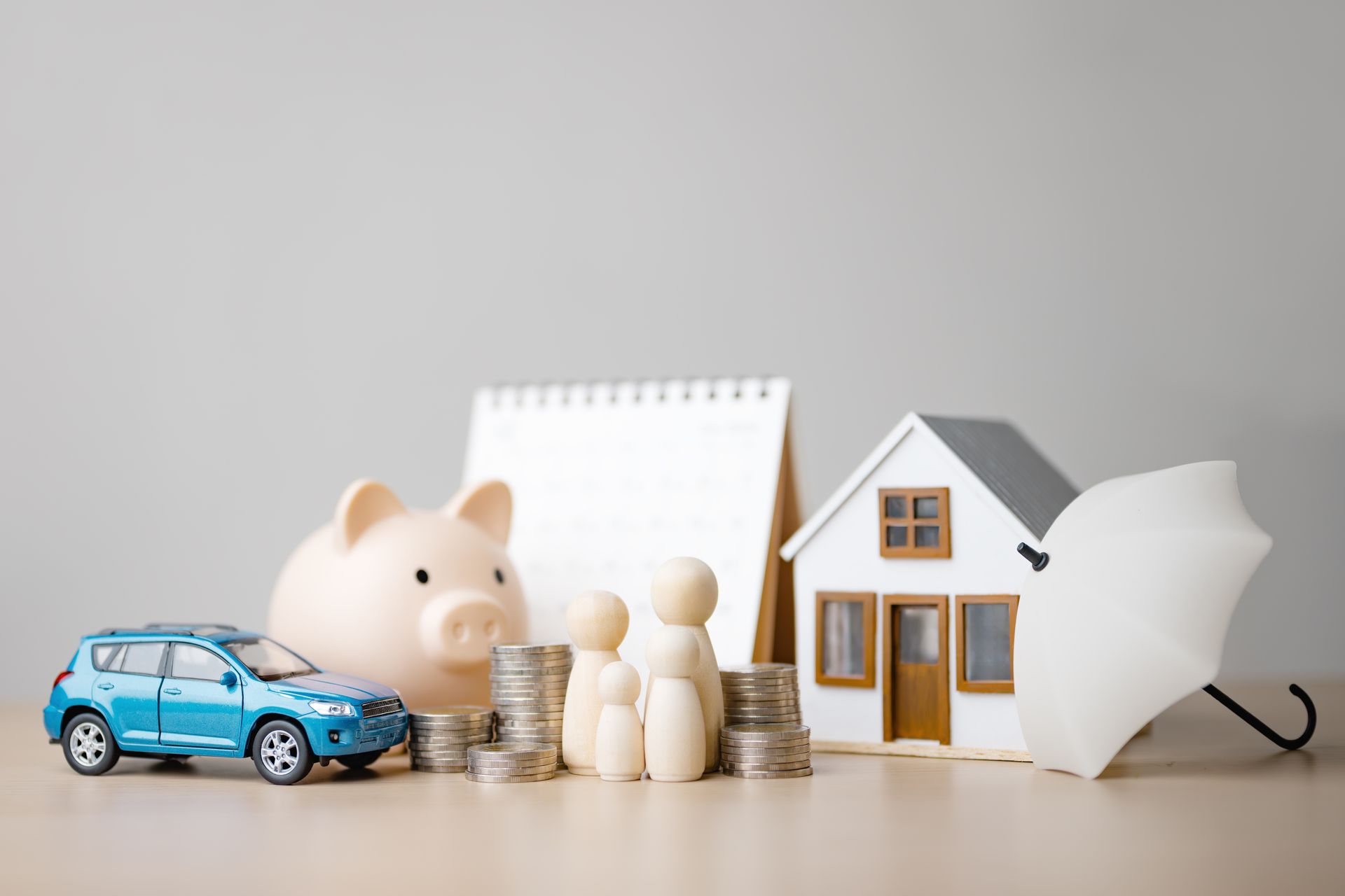 A blue toy car, piggy bank, stacked coins, wooden peg figures, a toy house, and an umbrella represent financial protection.
