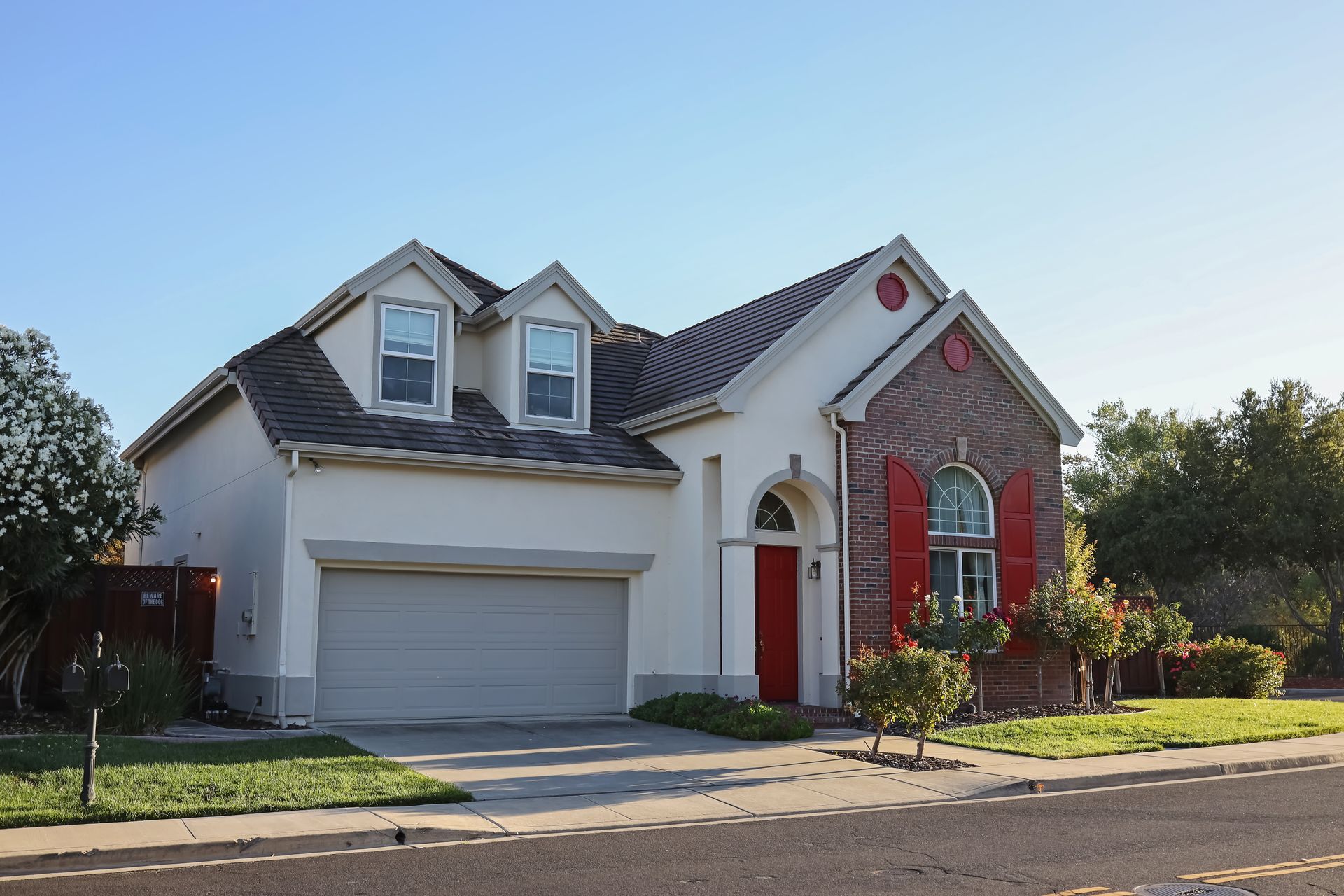 A light-colored house with a two-car garage, a red front door, and two dormer windows under a clear blue sky.