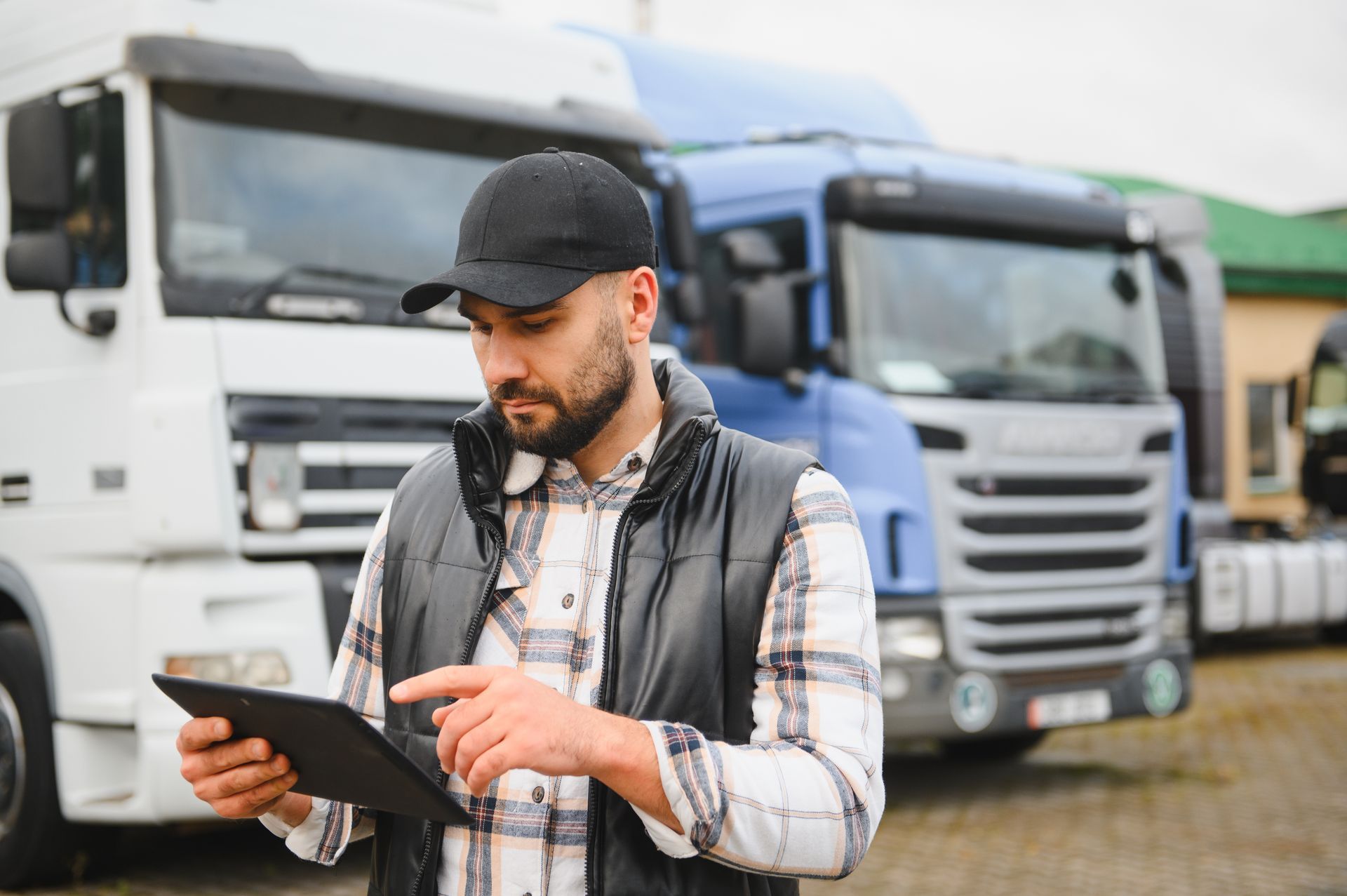 A person in a plaid shirt and vest uses a tablet while standing in a lot filled with semi-trucks.