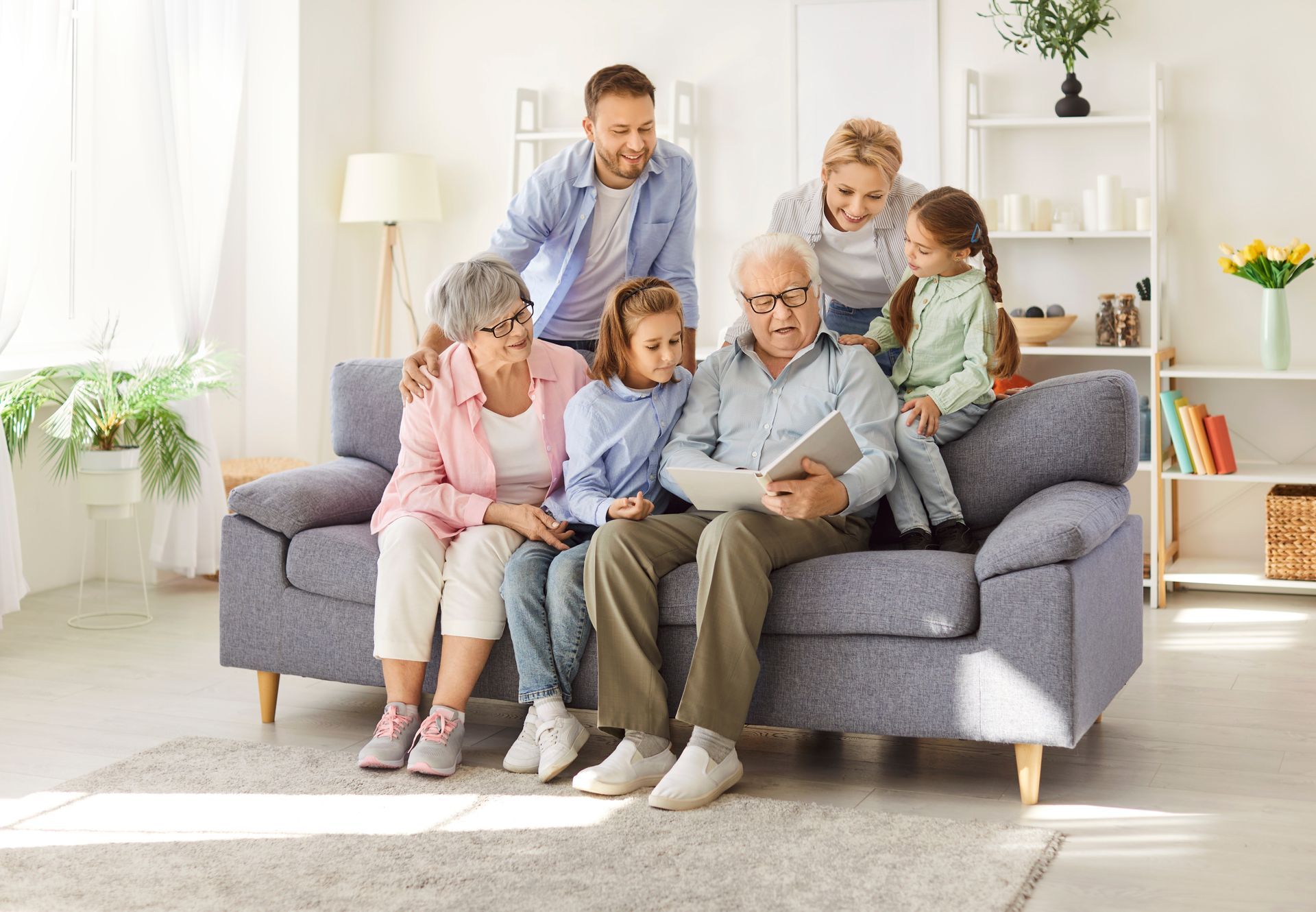 A family sitting on a sofa in a bright living room, gathered around an elderly person looking at a tablet together.