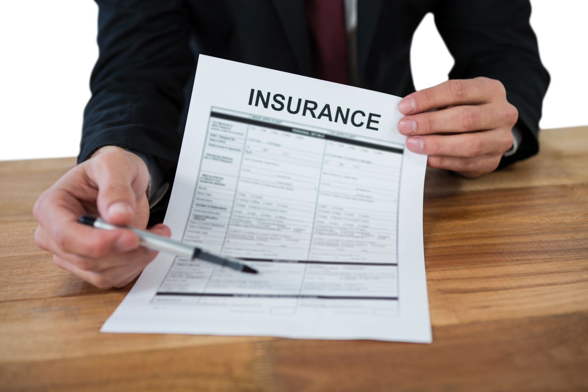 A person in a suit sits at a wooden desk holding a pen and pointing to an insurance document.