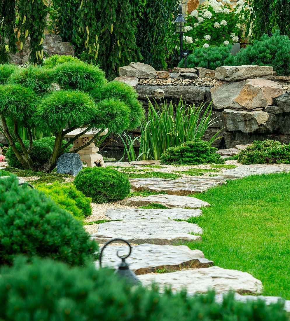 A stone path winds through a garden with manicured shrubs, green grass, and a rock wall feature in the background.