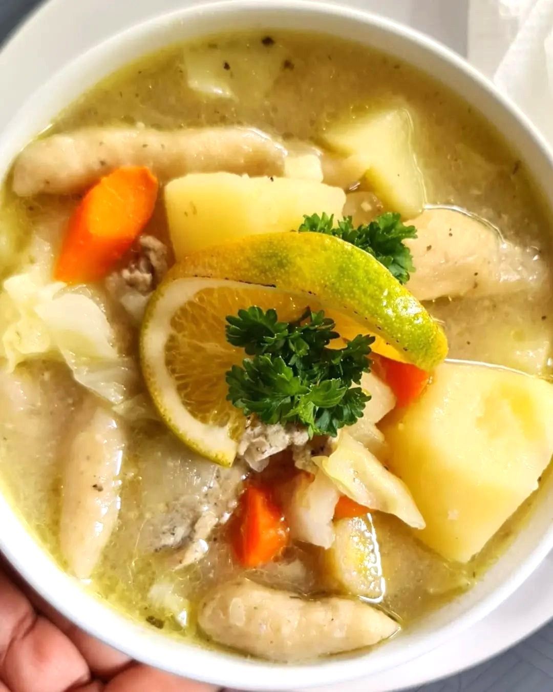 A bowl of soup with chicken , potatoes , carrots and lemon slices.