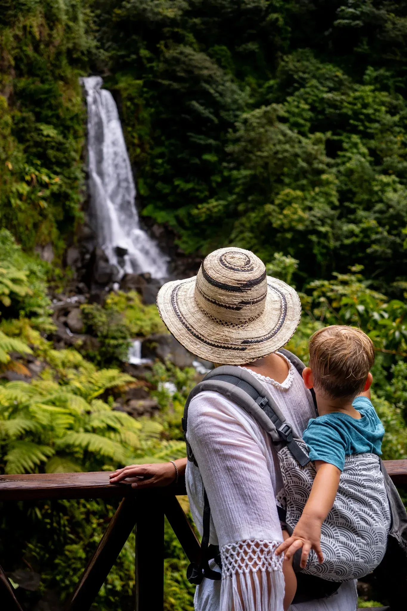 A woman with her baby attached to her back, looking at a waterfall.