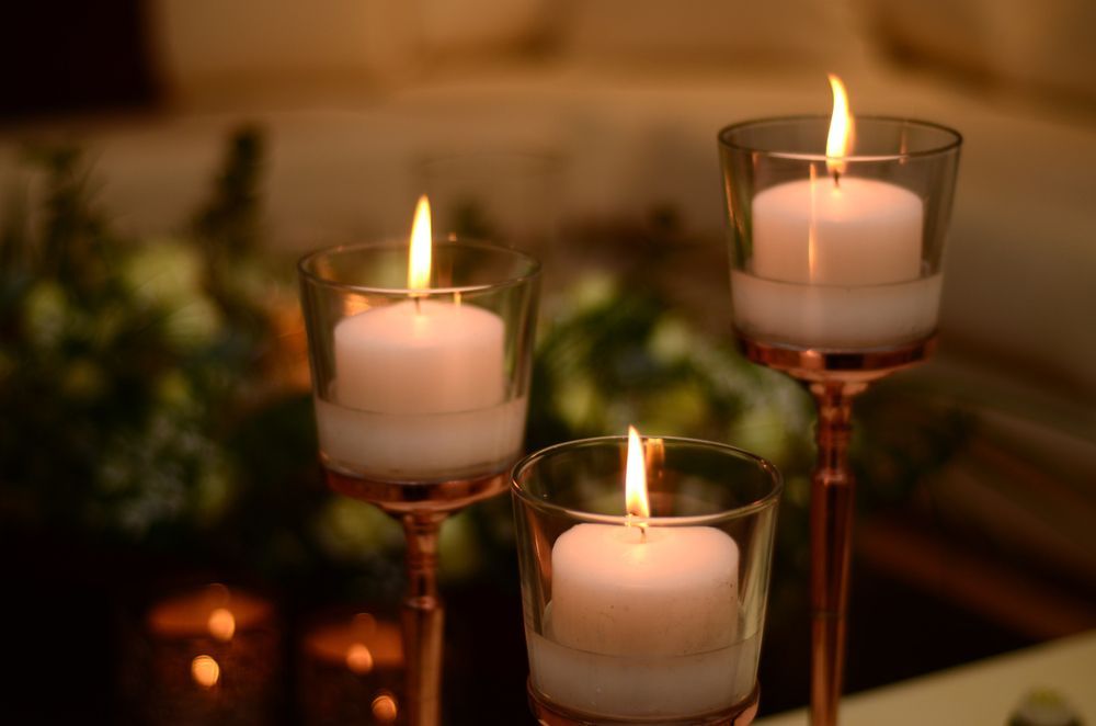 Three lit white candles in glass holders on stands, set against a blurred background of greenery.