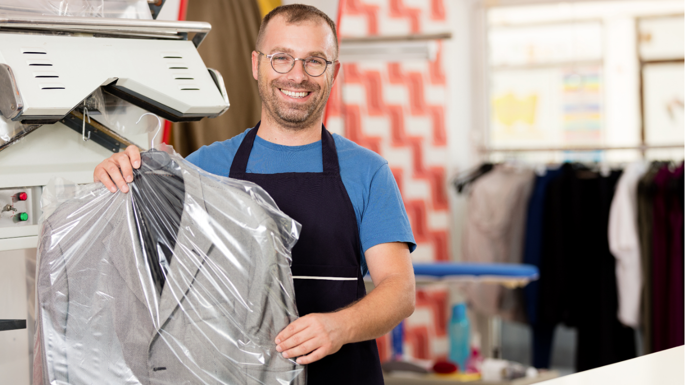 Man in apron holds a dry-cleaned suit in plastic wrap, smiling in a dry cleaning shop.