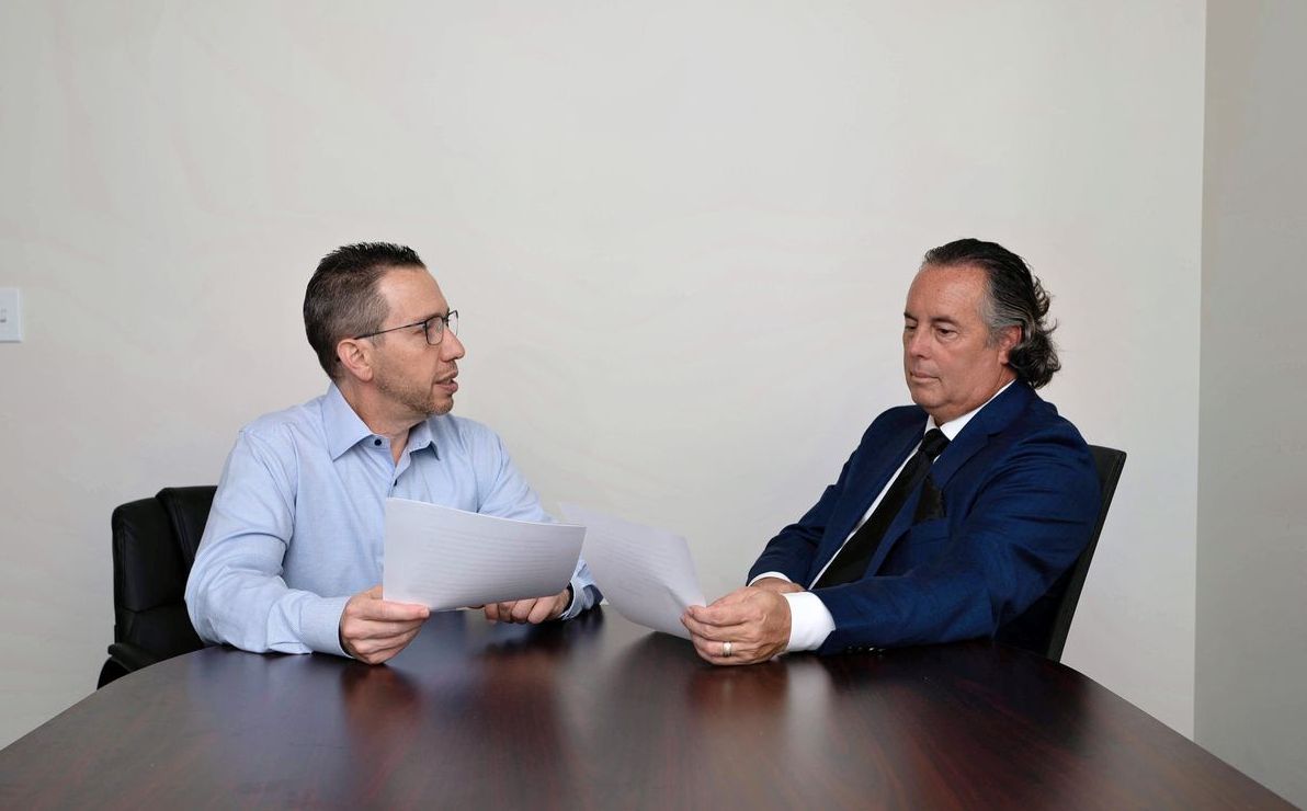 Two men are sitting at a table holding papers and talking to each other.