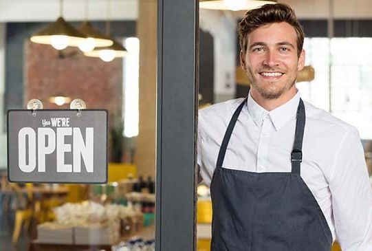 A man in an apron is standing in front of an open sign.