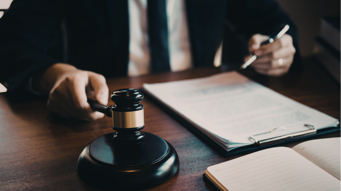 Gavel on desk in front of a person in a suit reviewing documents and holding a pen.