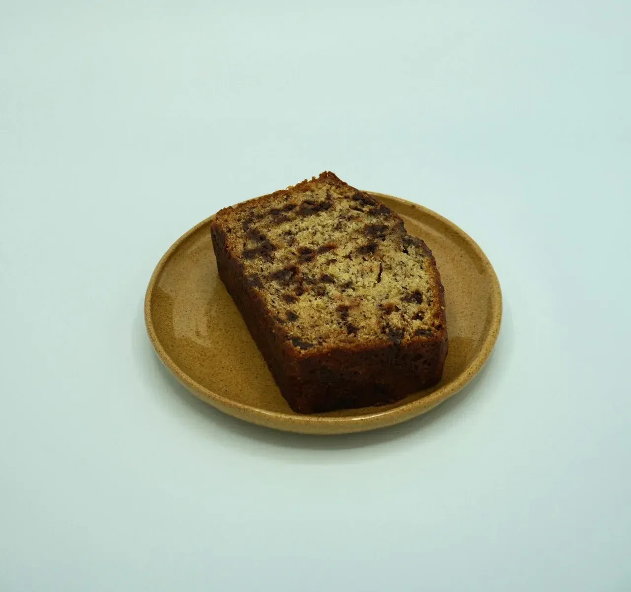 A single slice of banana bread with dark inclusions sits centered on a small, tan ceramic plate against a white background.