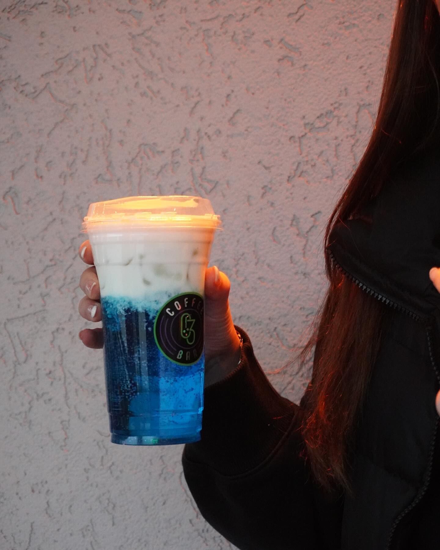 Person holding a layered blue and white drink in a clear plastic cup with a logo.