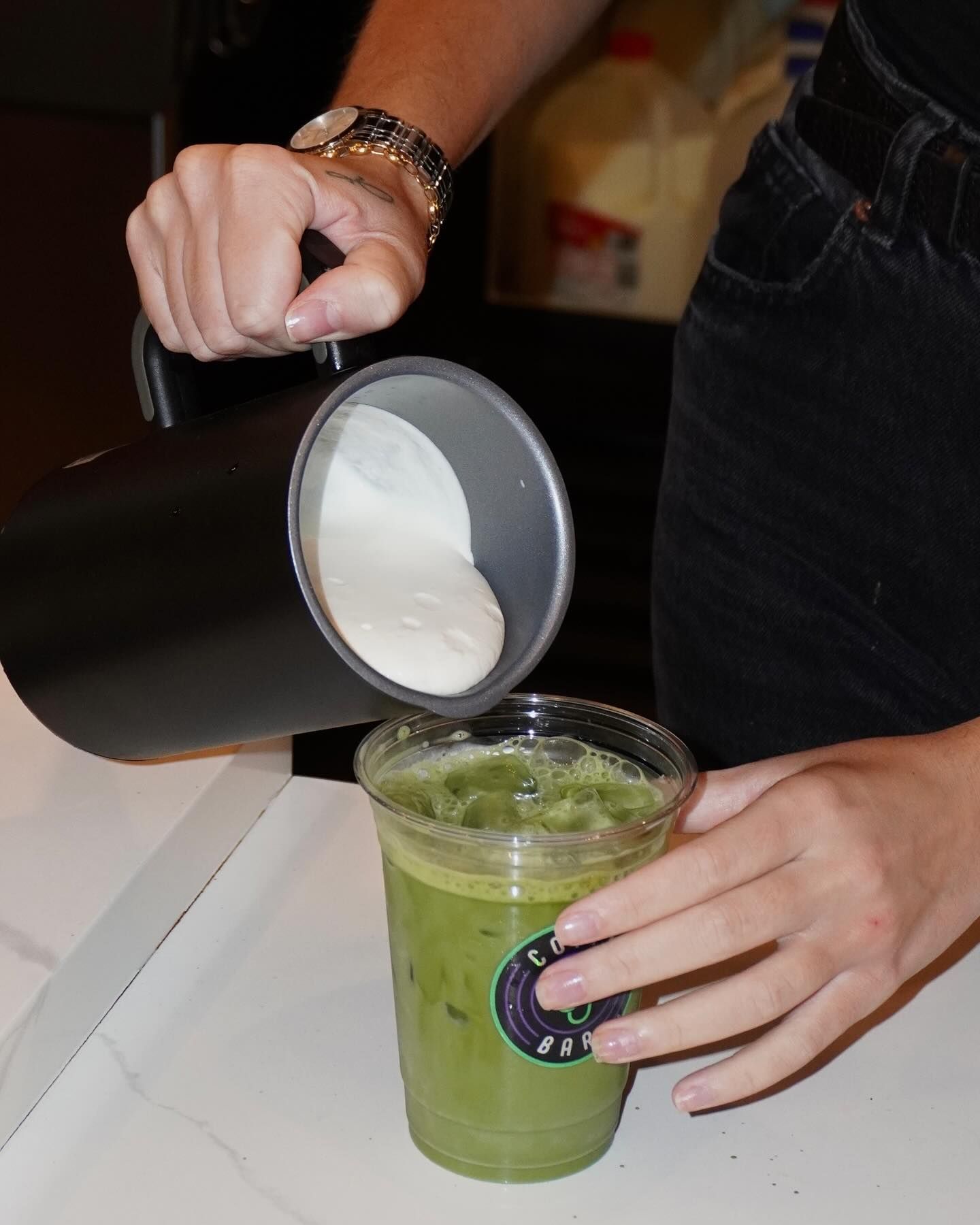 A barista pouring milk from a black pitcher into a green iced drink.