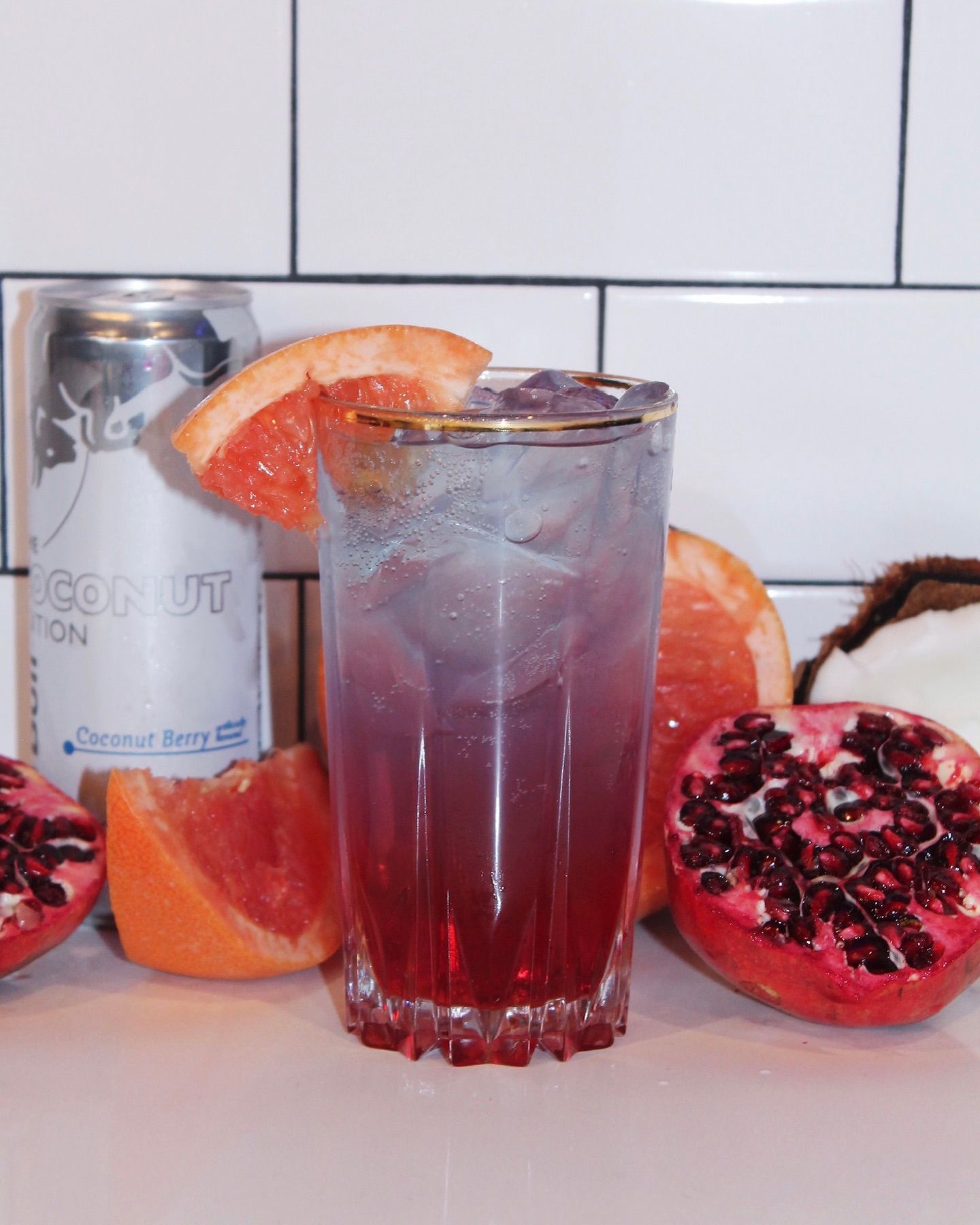 Tall cocktail with grapefruit and pomegranate, next to a coconut drink can and fruit.