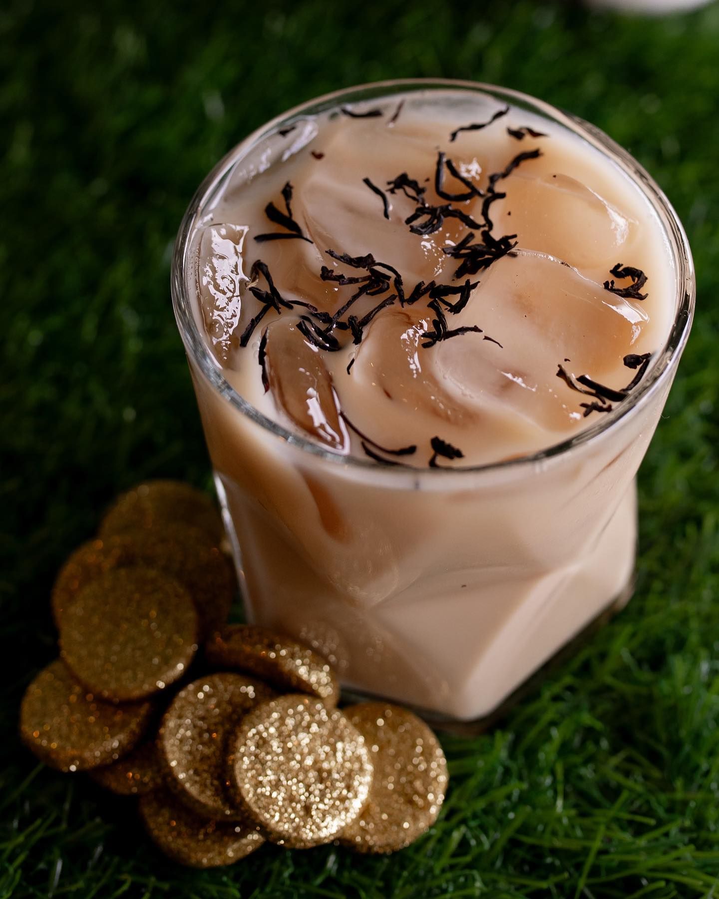 Iced drink with tea leaves on top, ice, gold coins, on a bed of green grass.