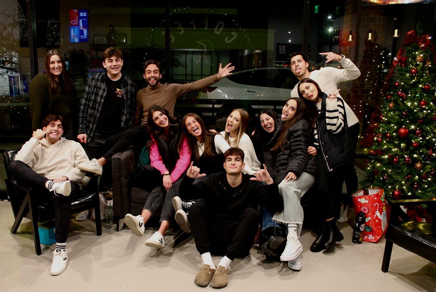 Group of friends smiling for a photo, near a Christmas tree. Many are sitting or leaning.