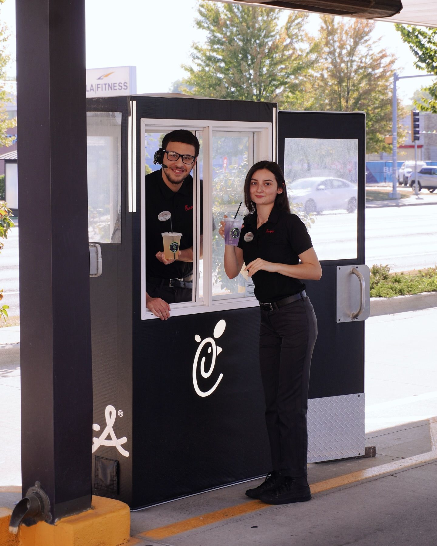 Two Chick-fil-A employees at a drive-thru window. One is smiling, holding a coffee cup, the other is holding a purple drink.