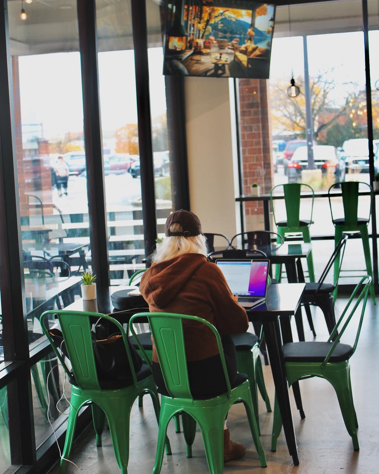 Person working on laptop at a table in a cafe, looking at screen. Green chairs, large windows, and brick wall visible.