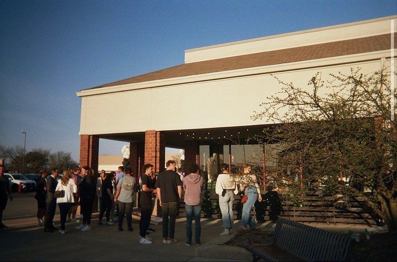 People gathered outside a building with a beige facade, brown trim, and glass doors. Evening light.