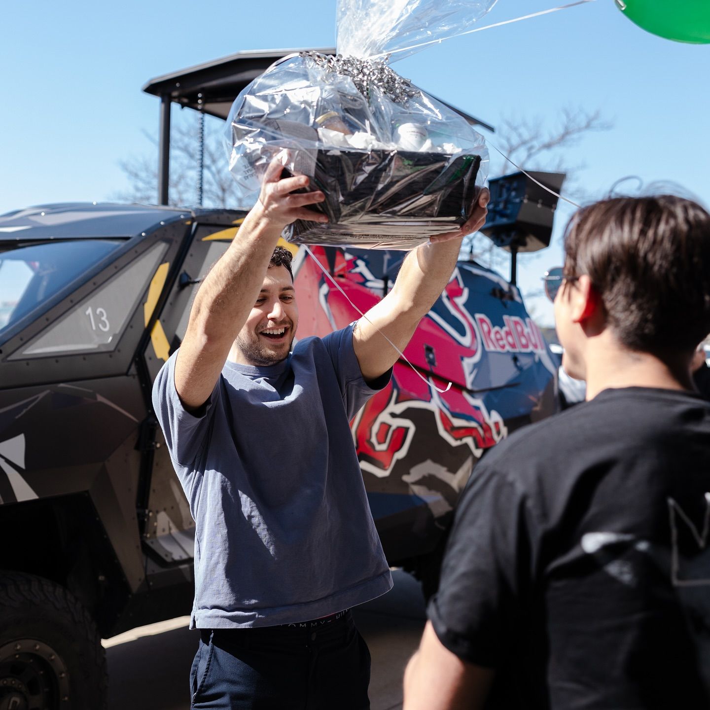 Man holding gift basket, smiling, near a black vehicle with a Red Bull logo; outdoors.
