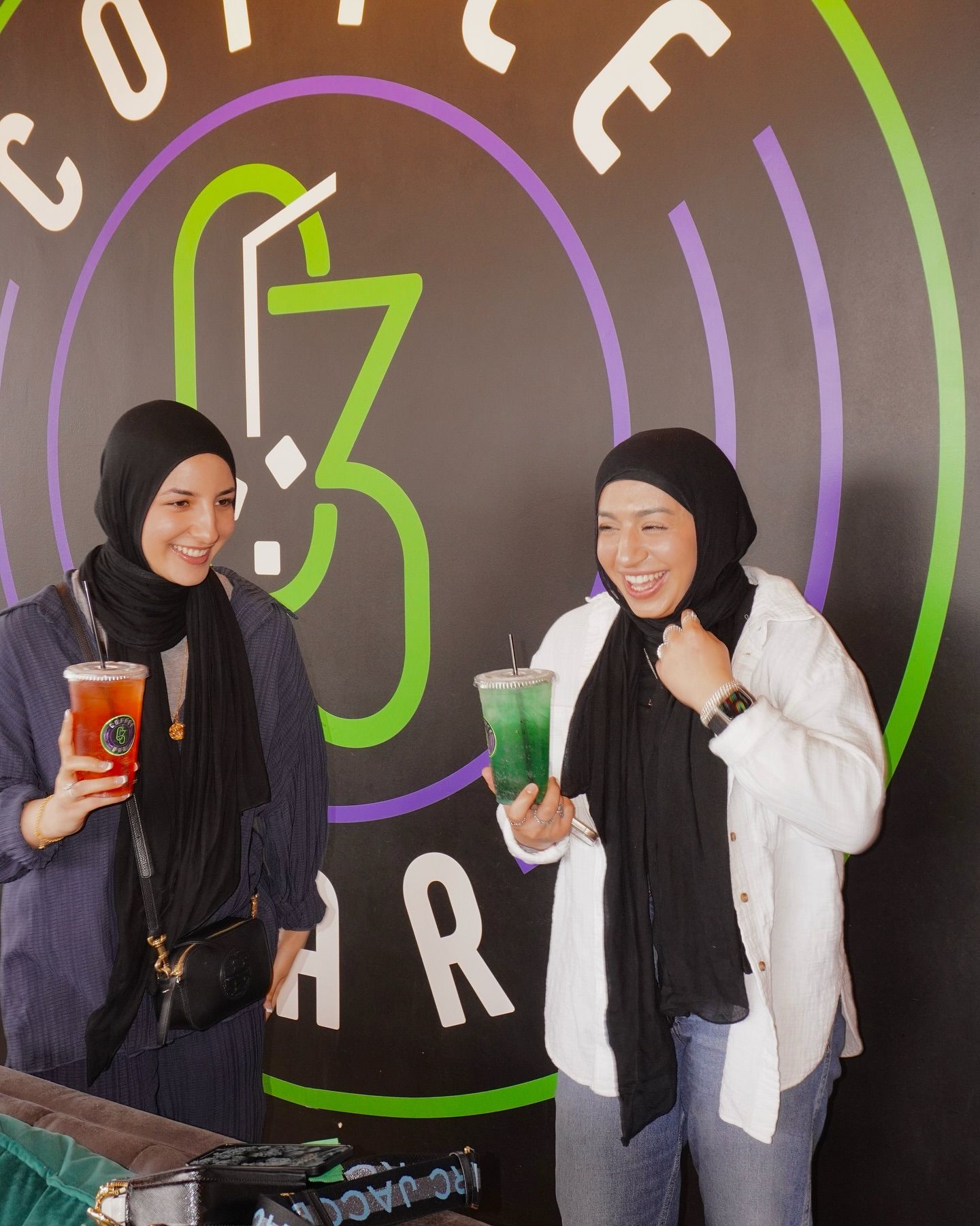 Two women in hijabs holding drinks, smiling in front of a coffee shop logo.