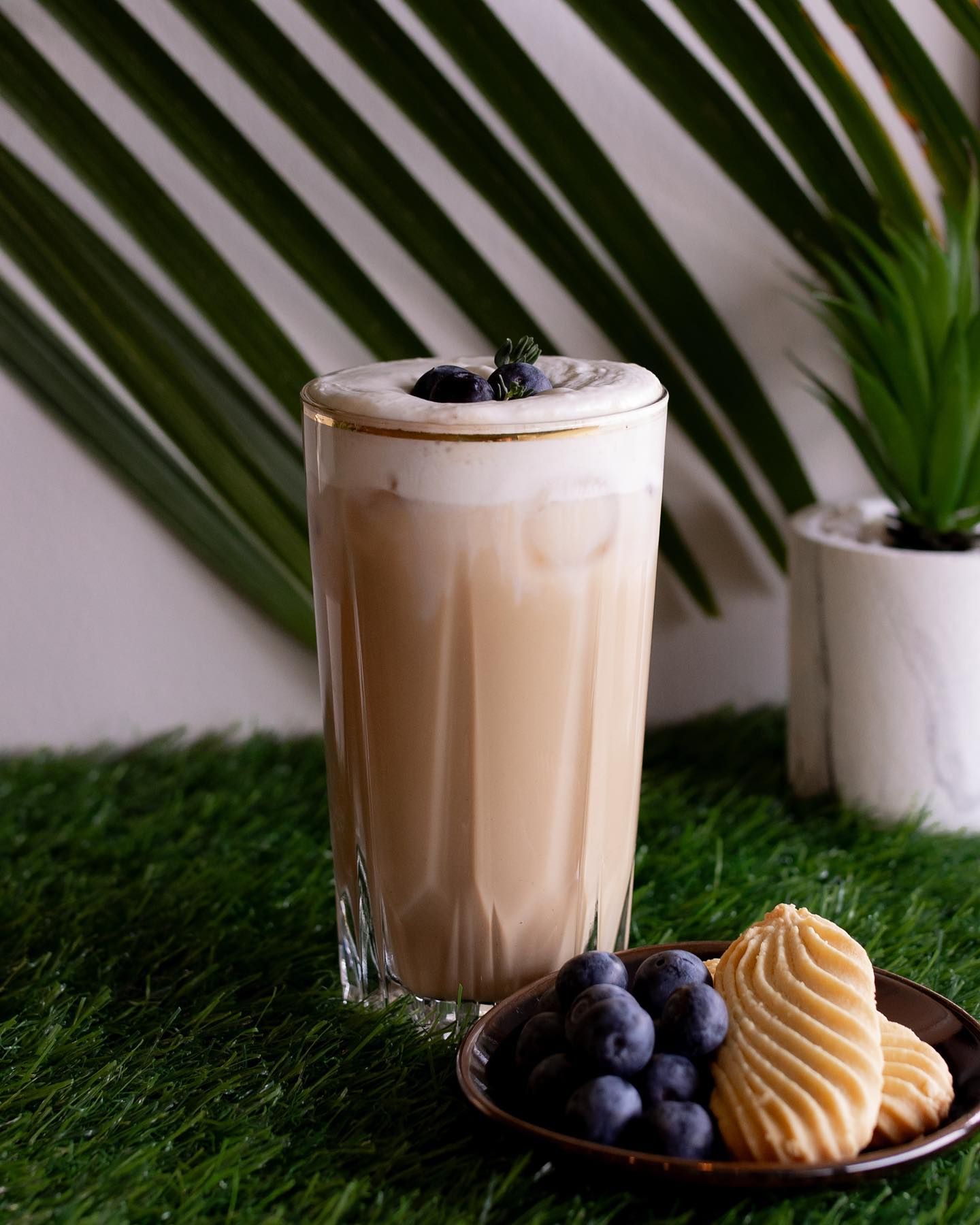 Tall glass of iced coffee topped with foam and blueberries, served with blueberries and cookies on a plate.