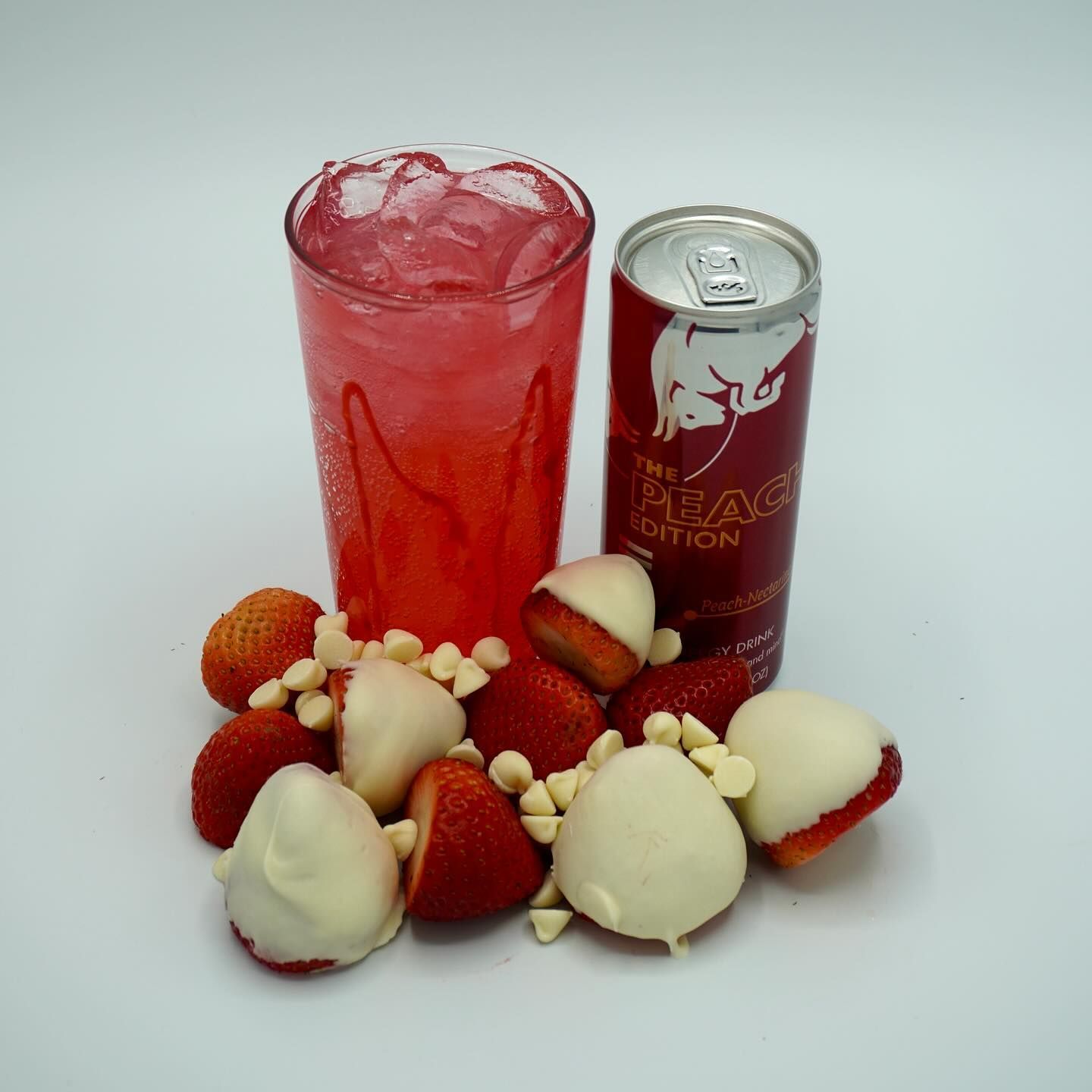 Glass of red drink with ice, Red Bull can, chocolate-covered and regular strawberries on white.