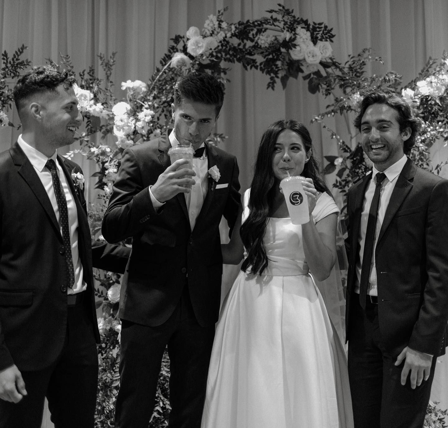 A bride and groom with groomsmen drinking from cups in front of a floral arch; black and white photo.