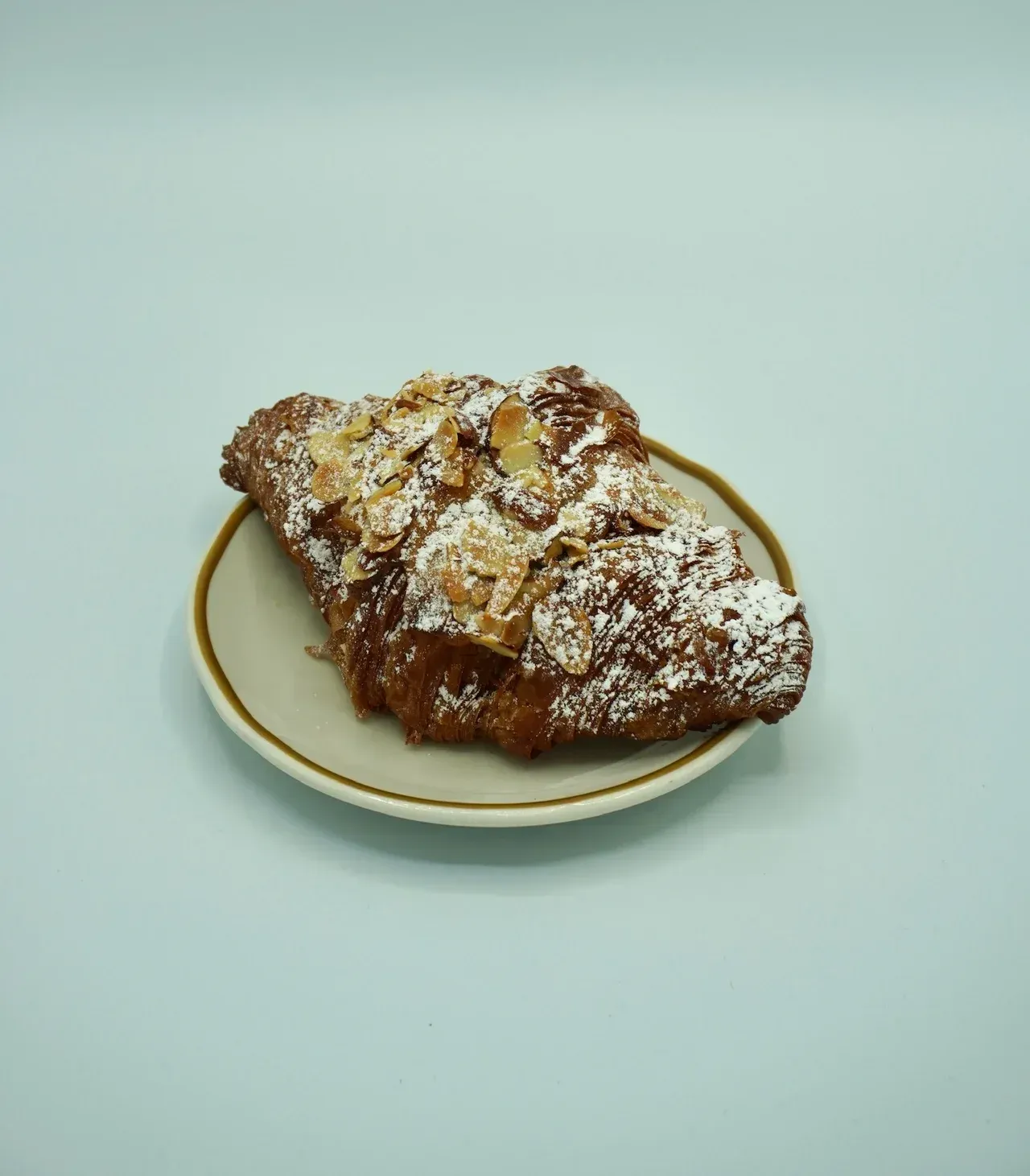 An almond croissant topped with powdered sugar, sitting on a small plate against a light blue background.