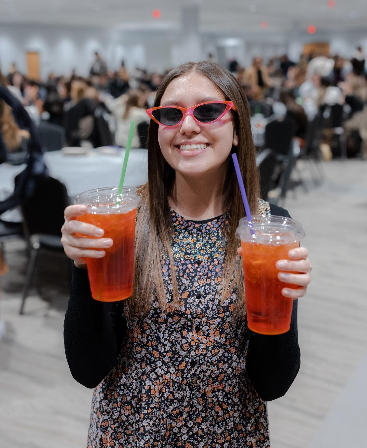 Woman holding two drinks with straws, wearing pink sunglasses, smiling in a large room with people.